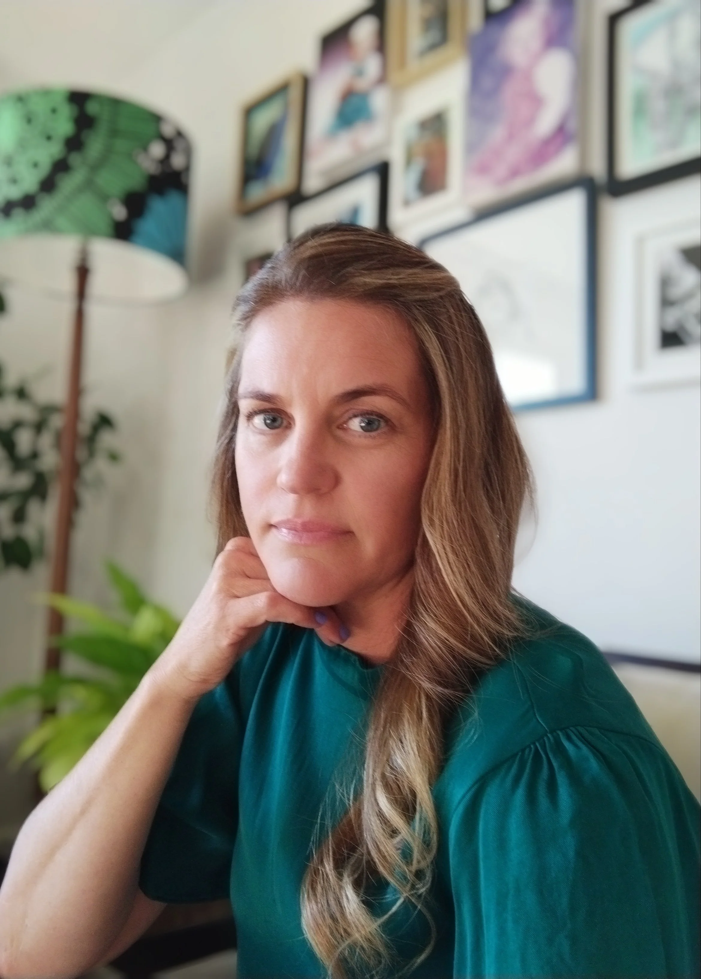 A woman with long light brown hair and blue eyes, wearing a teal top, resting her chin on her hand, sitting indoors with a gallery wall and a plant in the background.