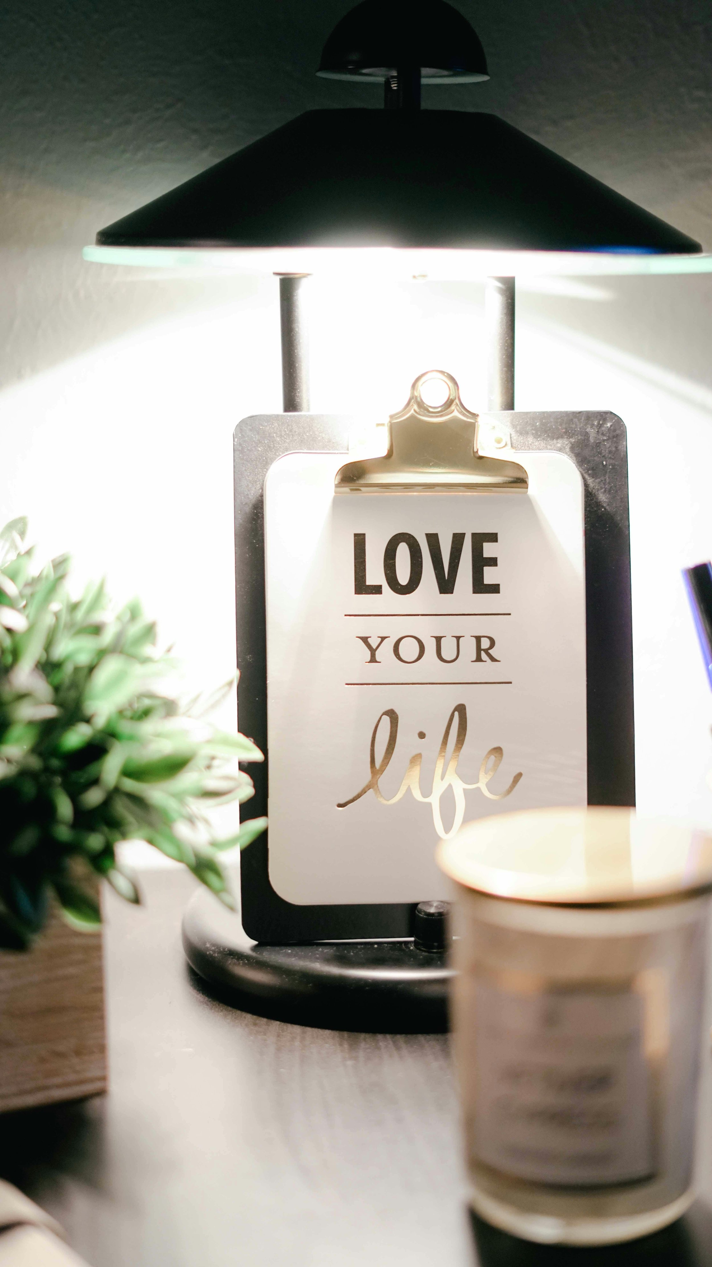 A bedside lamp illuminating a sign that says, 'Love Your Life,' with a small plant on the left and a lit candle on the right.