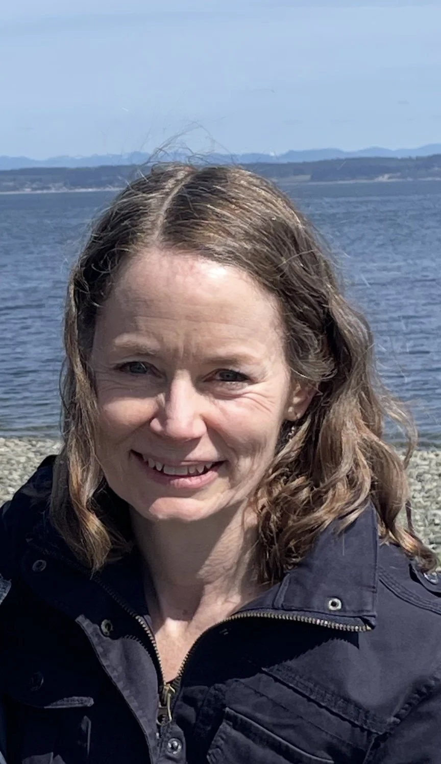 A woman smiling outdoors with a body of water and mountains in the background.