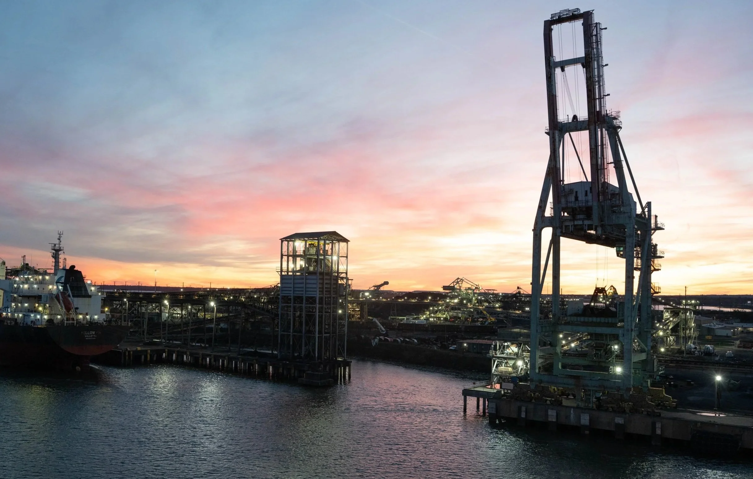 Shipyard at sunset with large industrial cranes and ships docked in the water.