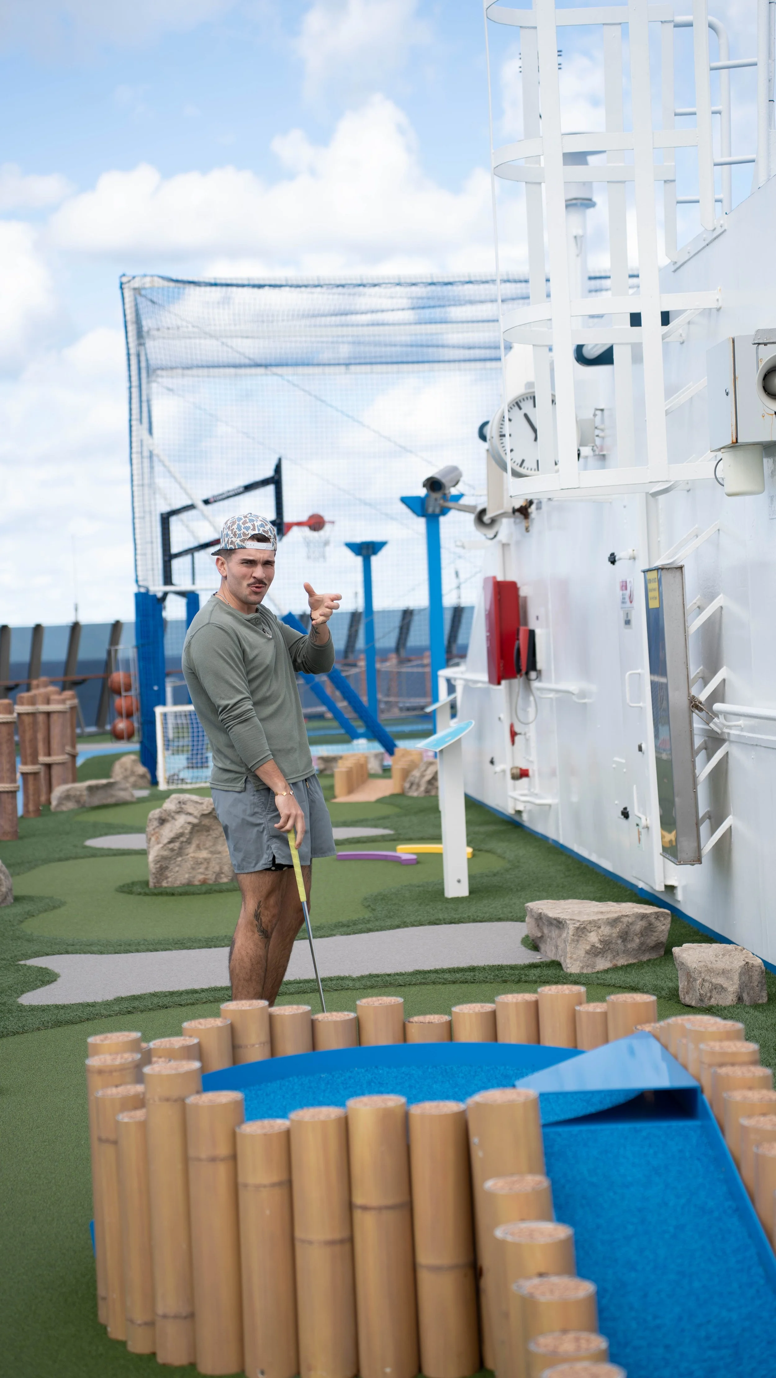 Young man at minigolf course holding putter, wearing casual clothes and a cap. Miniature golf obstacle in the foreground, artificial turf, playground equipment, and a sports court with basketball hoops in the background under a partly cloudy sky.
