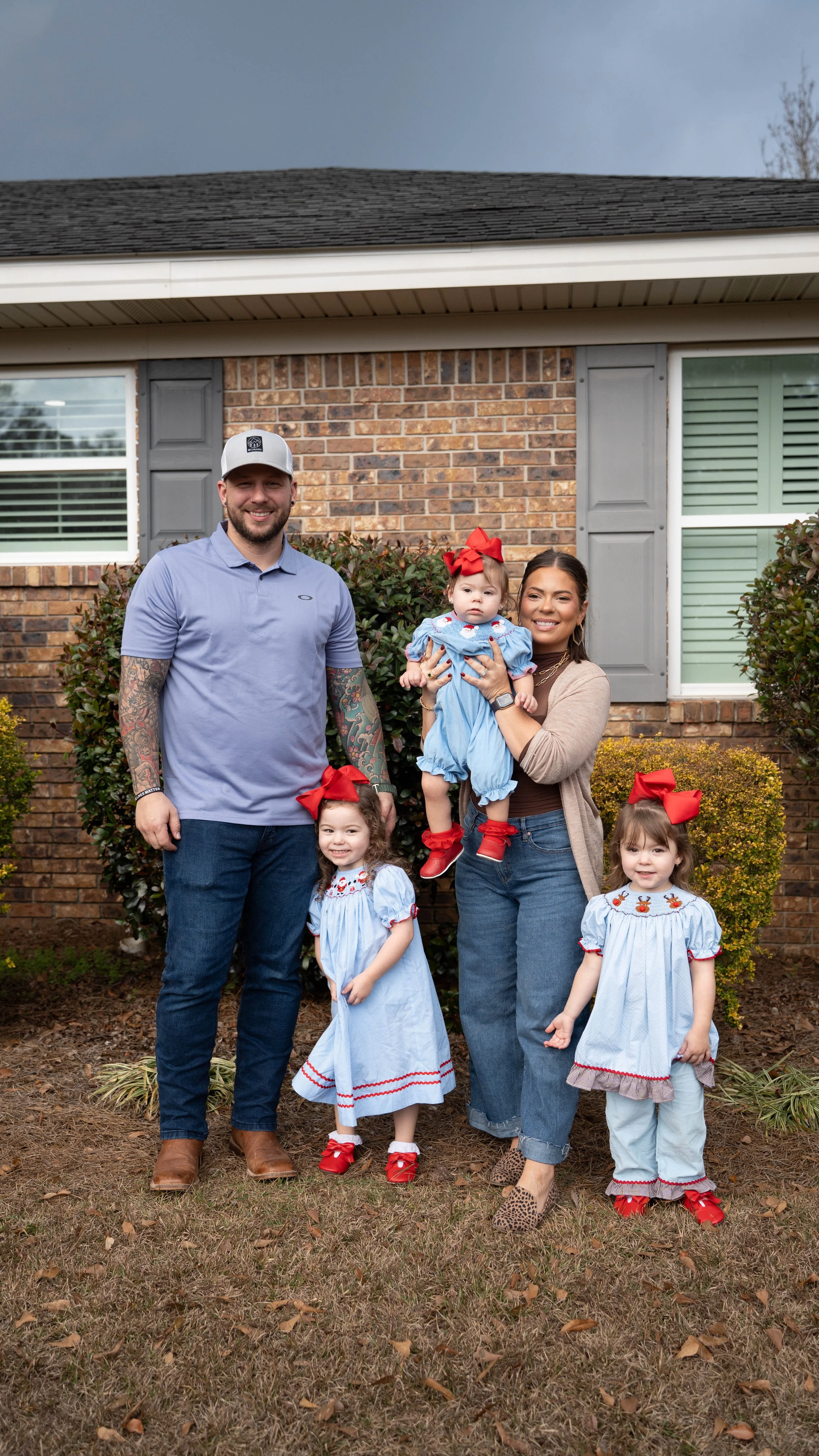 A family of five standing outdoors in front of a brick house with gray shutters. The father has tattoos on his arms and is wearing a light blue polo, jeans, and a white cap. The mother, holding a young girl, is wearing a beige cardigan, jeans, and leopard print shoes. Two young girls are in front, all dressed in matching blue outfits with red bows and shoes. The family is smiling, and the background includes shrubs and a cloudy sky.