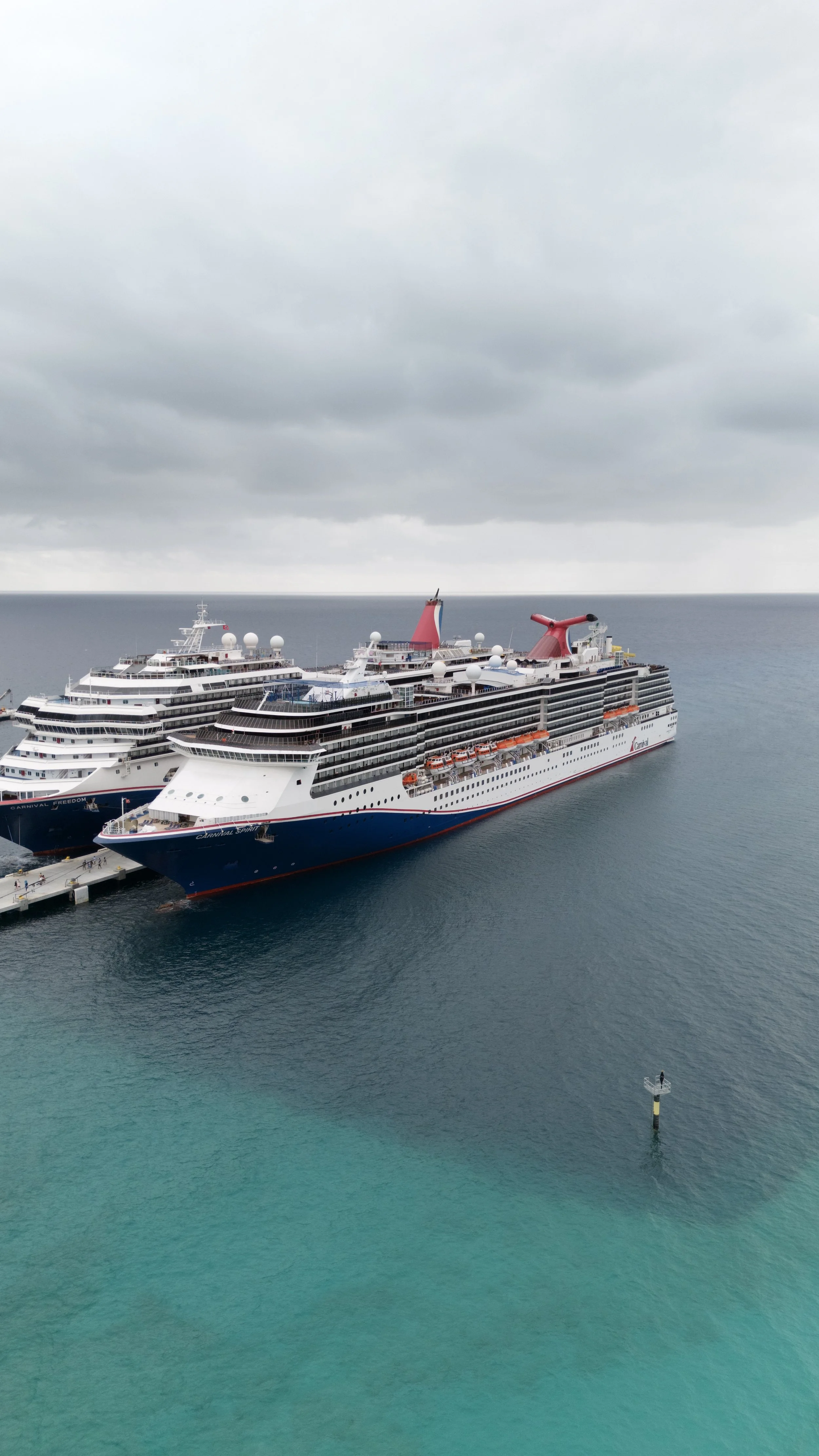 Large cruise ship docked at a pier on a cloudy day with calm turquoise water in the foreground.