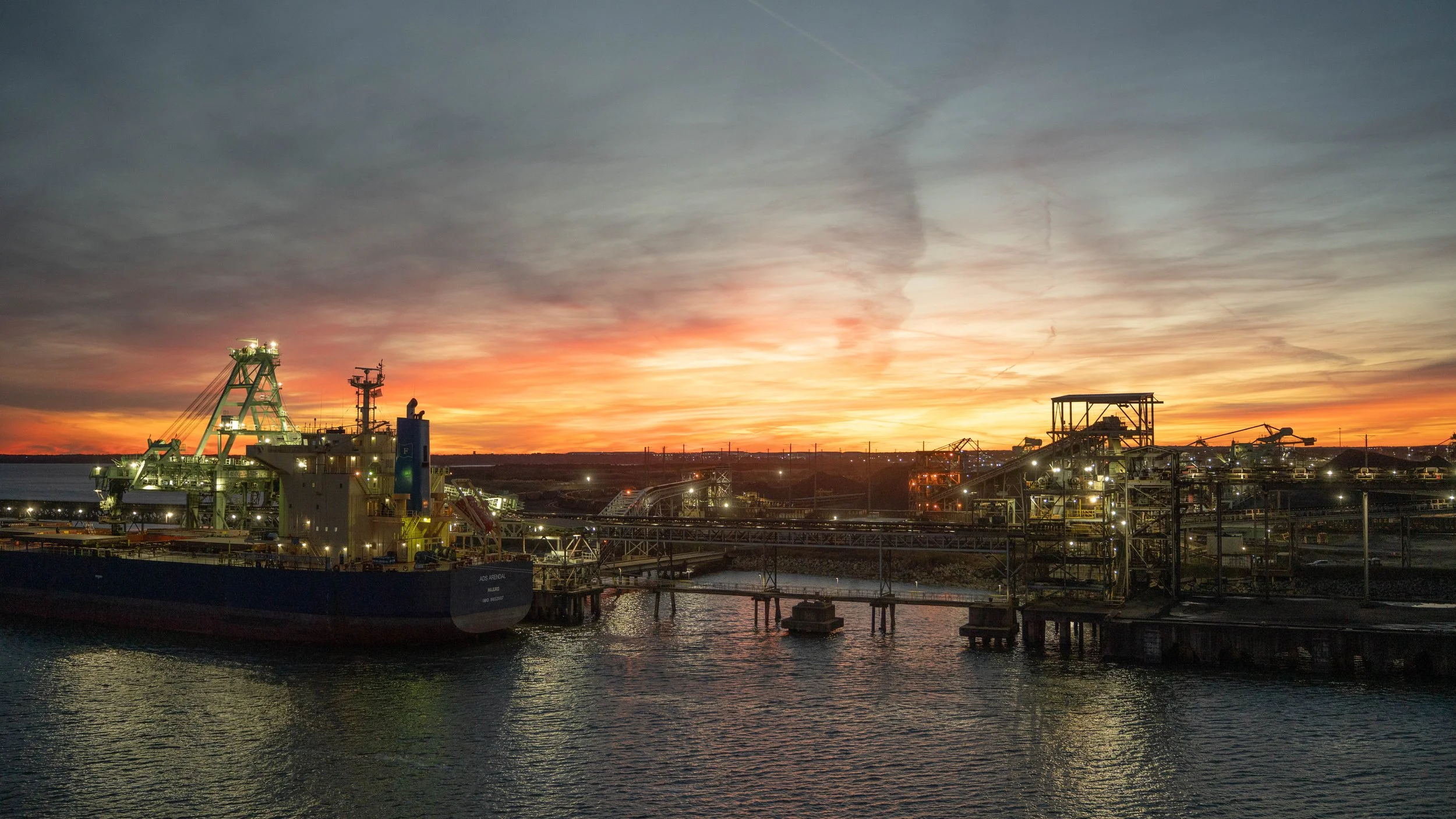 Industrial oil or gas refinery at sunset with water in the foreground and a colorful sky.