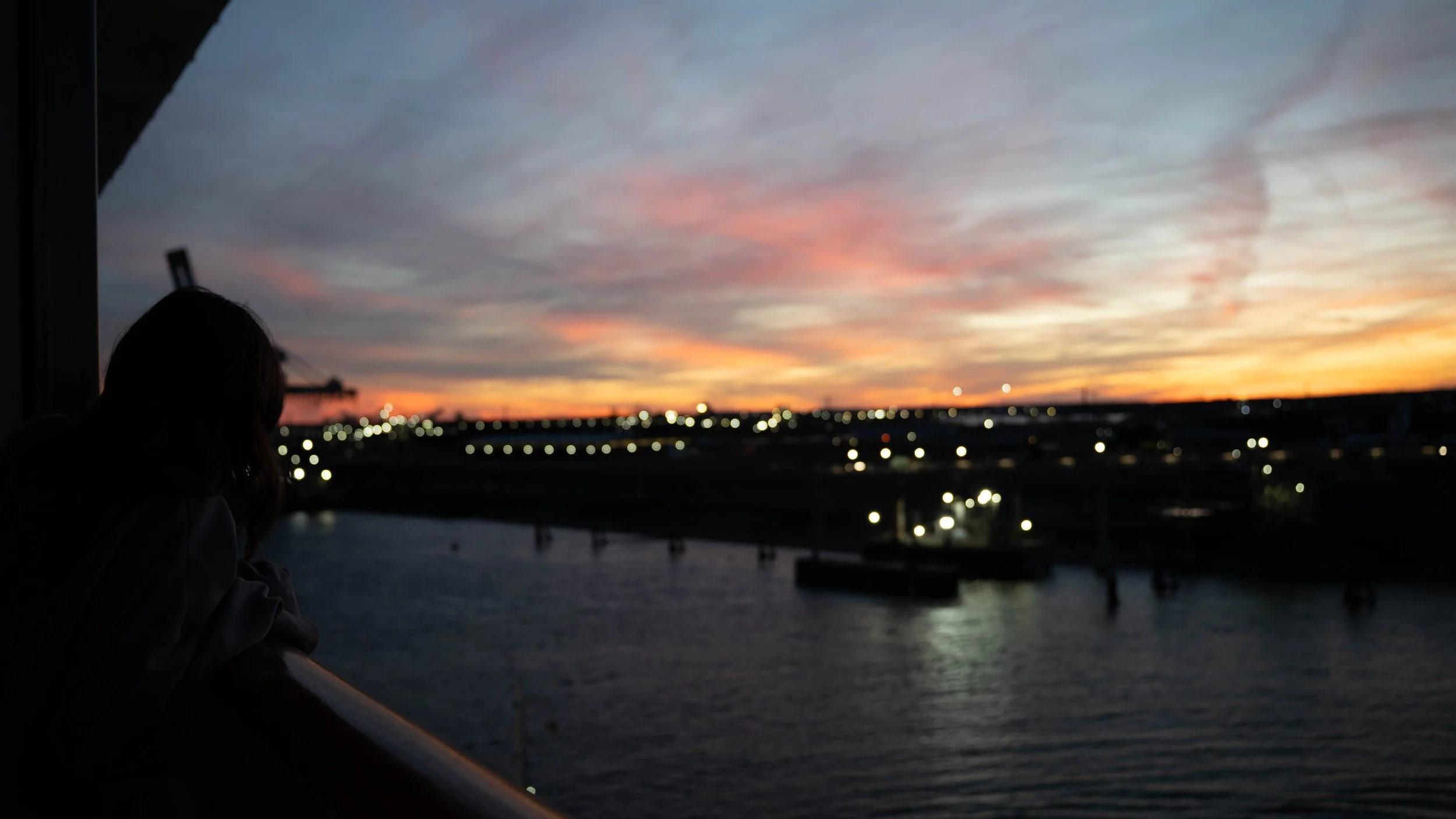 Person looking out over a river at sunset, with a colorful sky and city lights in the distance.
