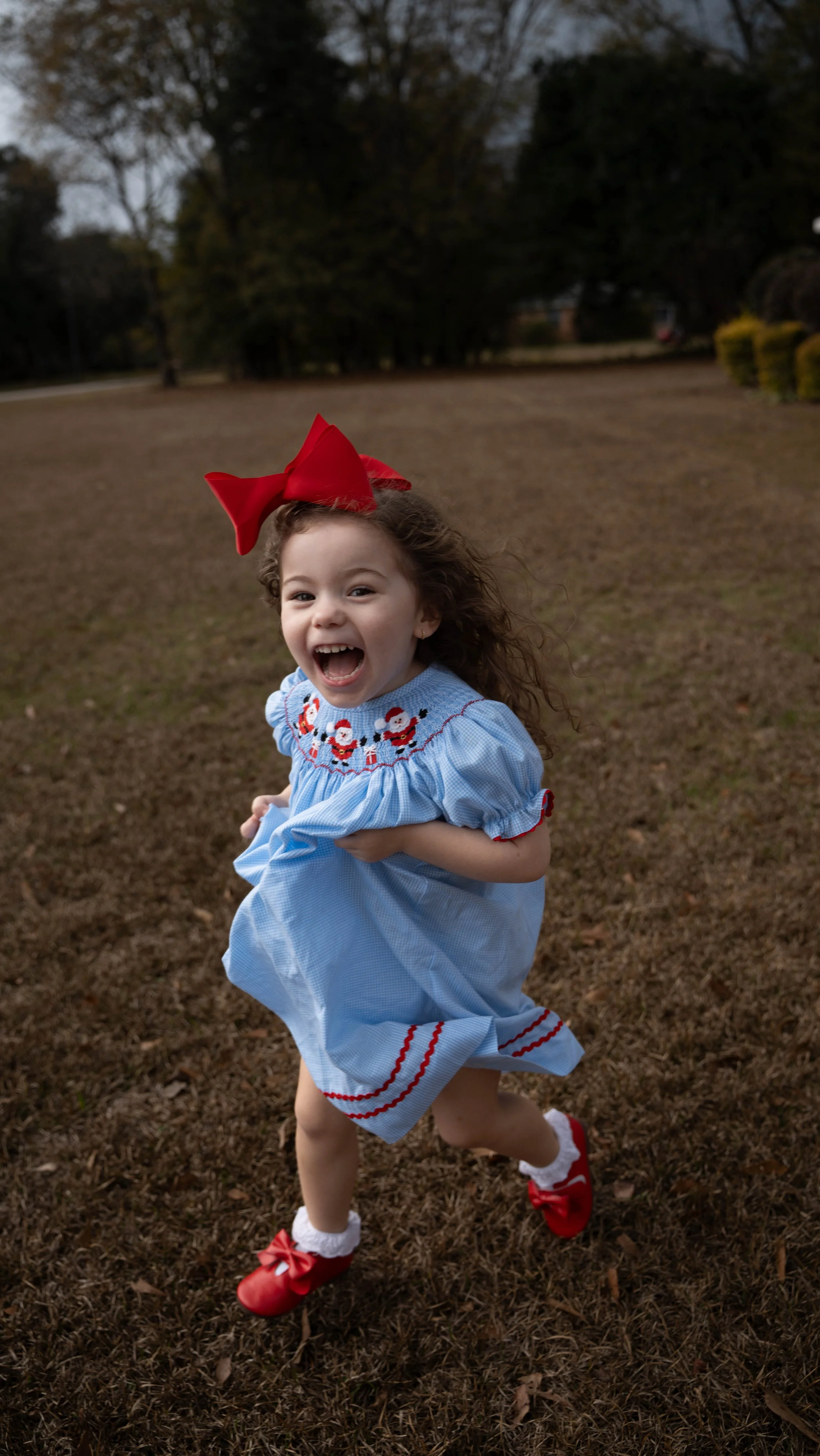 A young girl running outdoors in a grassy area, wearing a Christmas-themed dress with Santa Claus patterns, a big red bow in her hair, and red shoes with bows.