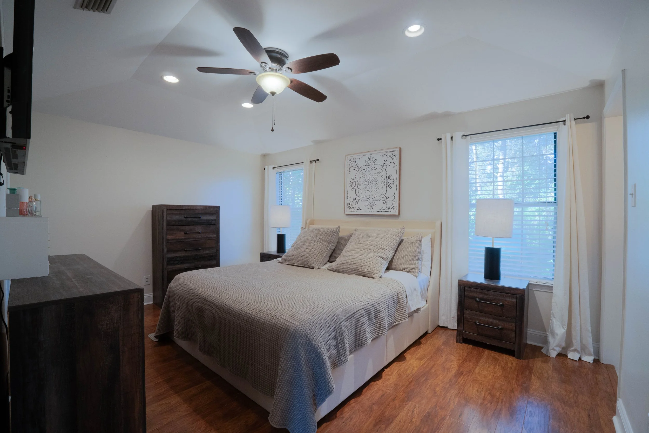  A bedroom with a bed, two nightstands with lamps, a dresser, a ceiling fan, and two windows with white curtains, hardwood floors. Mobile, Alabama