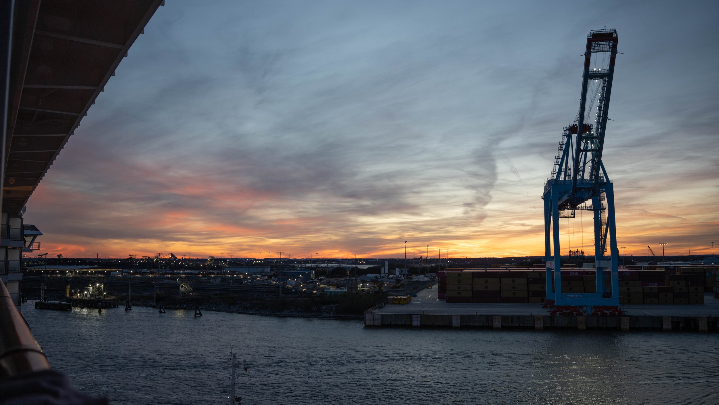View of a port or industrial waterfront at sunset with a large blue cargo crane, shipping containers, and a waterway in the foreground.