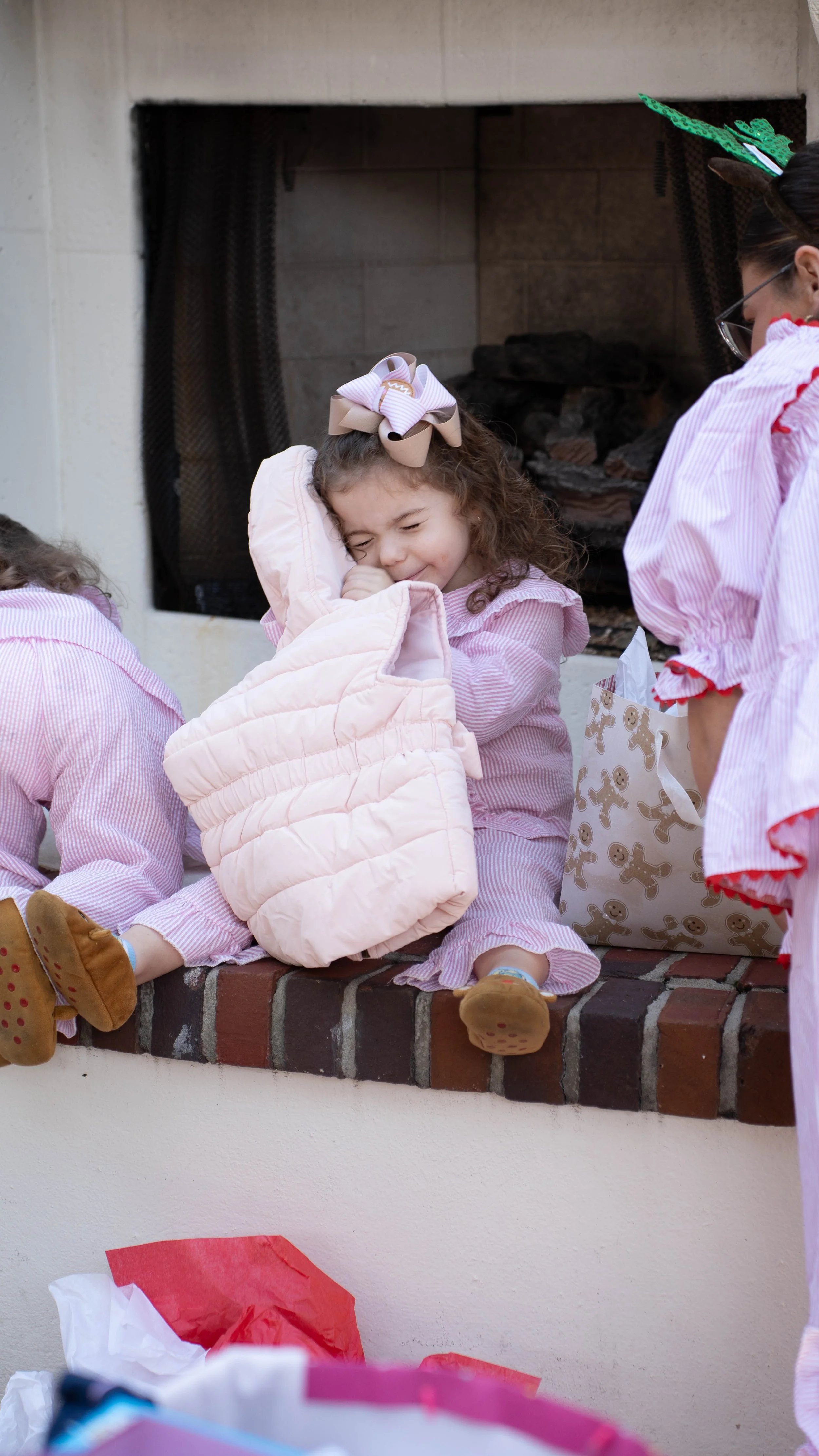 A young girl sitting on a brick fireplace, hugging a pink quilted blanket with her eyes closed and a smile on her face, wearing pink pajamas and a bow in her hair, during a holiday celebration.
