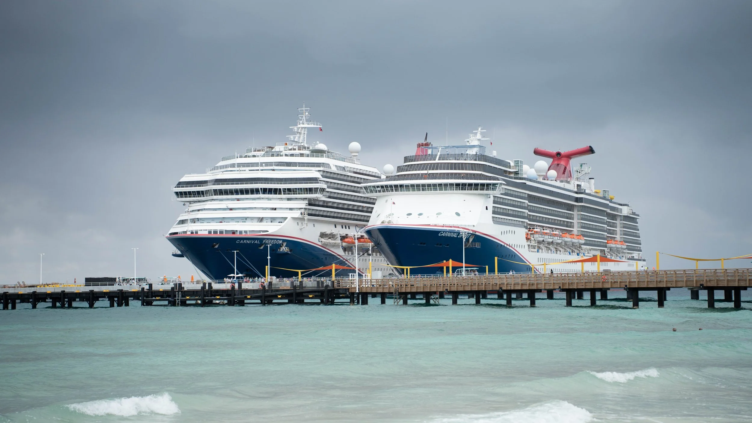 Two large cruise ships docked at a pier on a cloudy day, with calm water in the foreground.