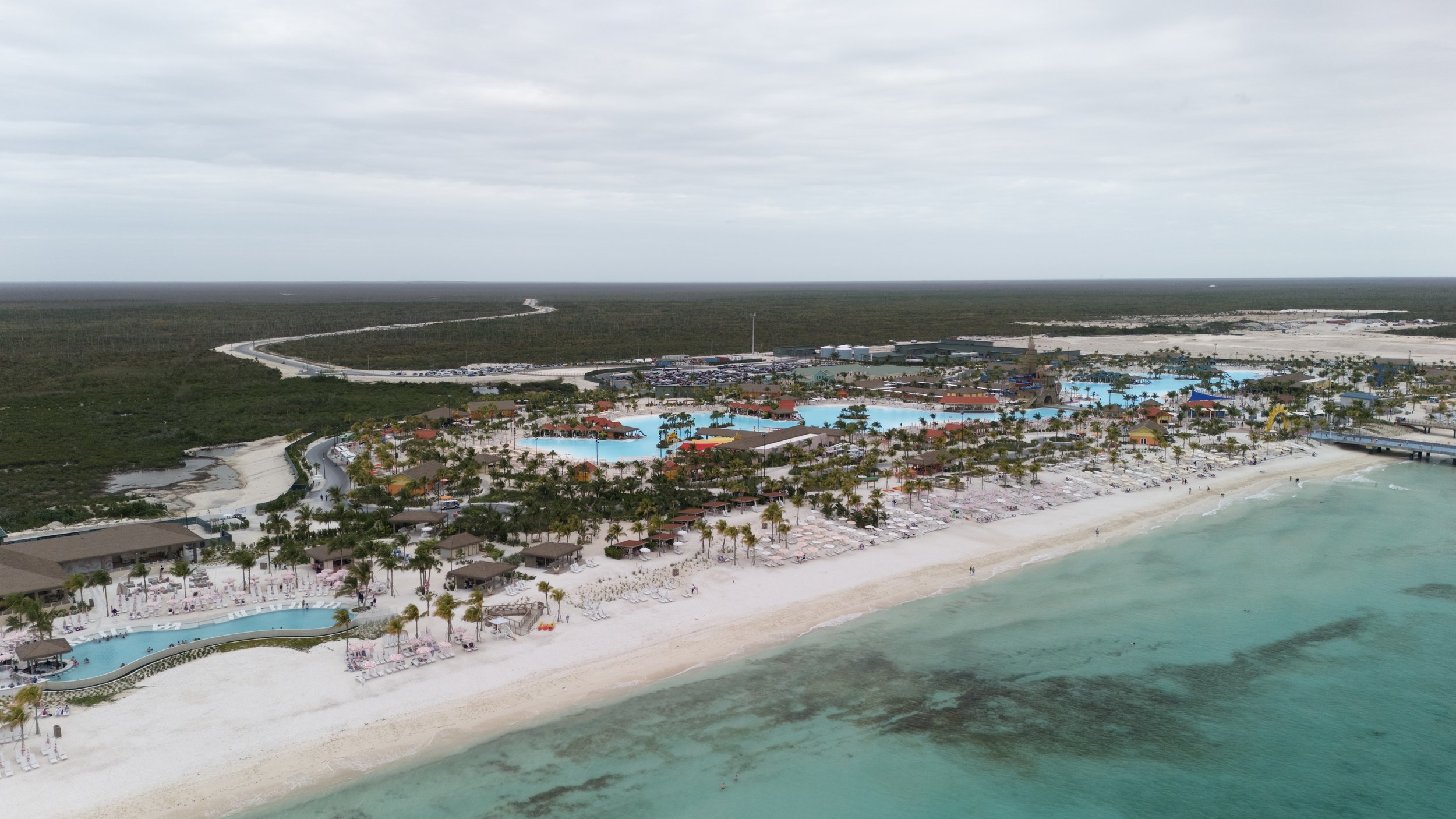 An aerial view of an all-inclusive beach resort with multiple pools, sun loungers, umbrellas, palm trees, and a sandy beach along turquoise water, surrounded by green wilderness.
