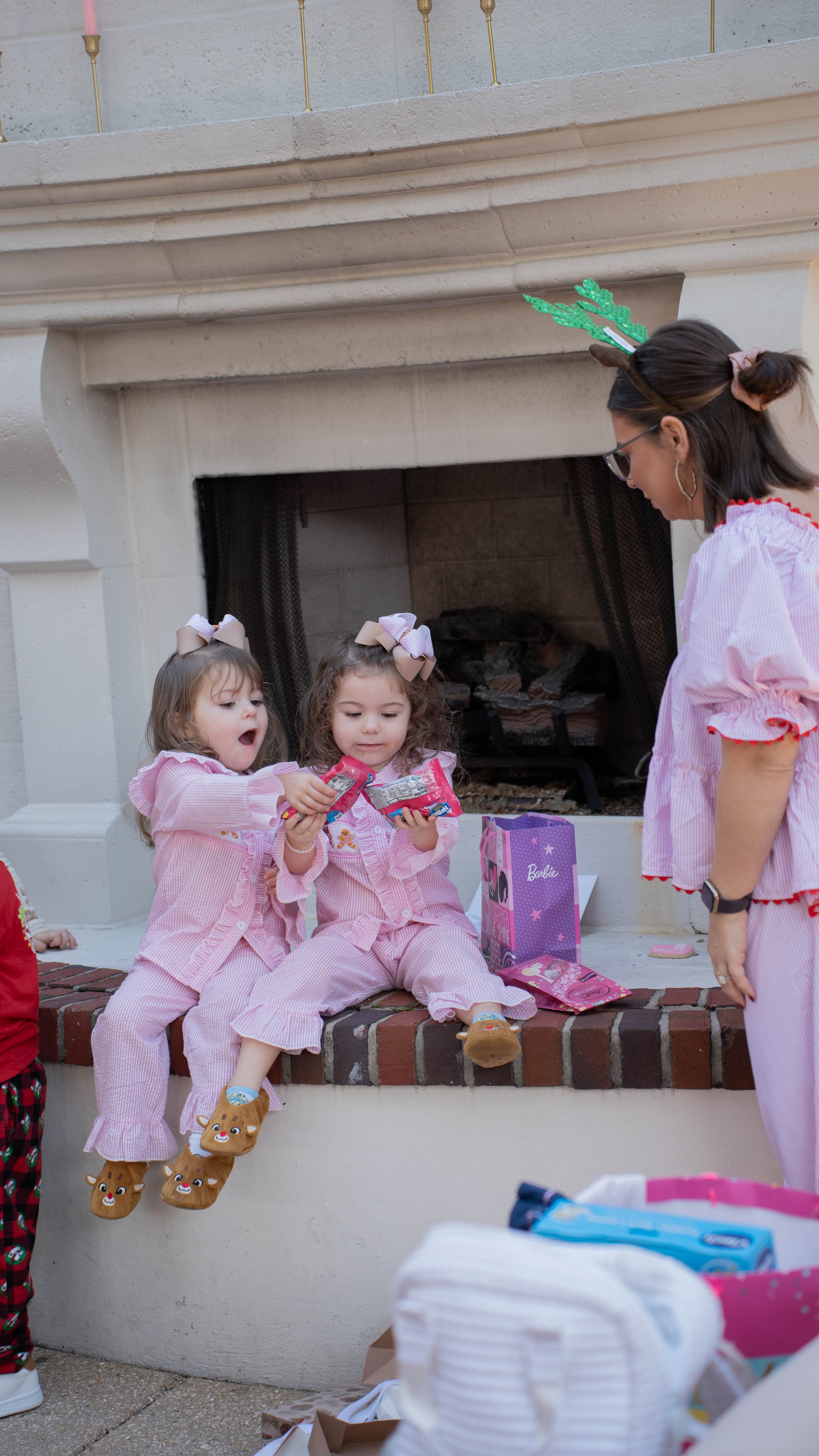 Two young girls in pink pajamas sitting on a brick ledge in front of a fireplace, opening pink toy packages, with an older woman in holiday attire talking to them. One girl is wearing reindeer slippers, and the woman is wearing reindeer antlers.
