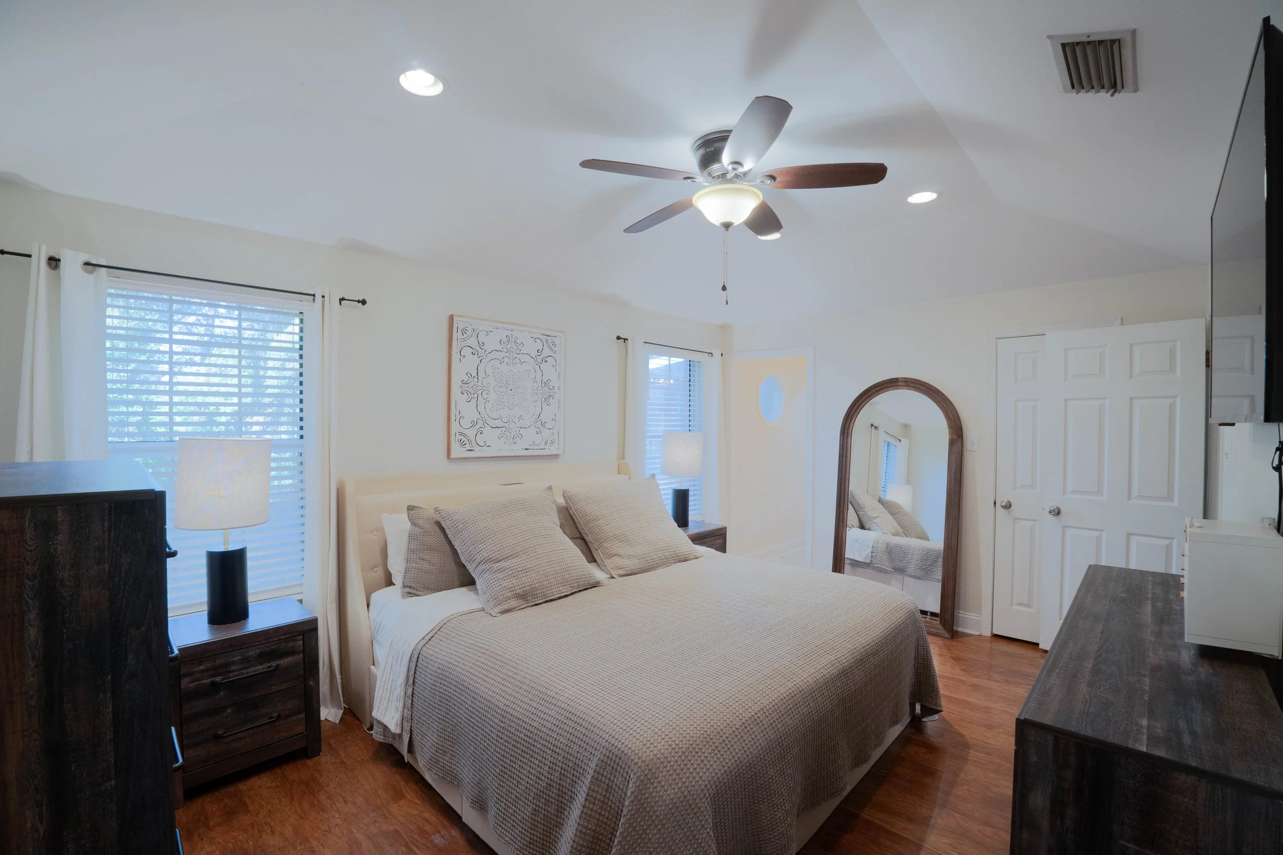 A bedroom with a large bed with beige bedding and pillows, dark wood nightstands with lamps, a mirror, a ceiling fan, windows with blinds and curtains, wooden flooring, and a closed closet door. Mobile,AL