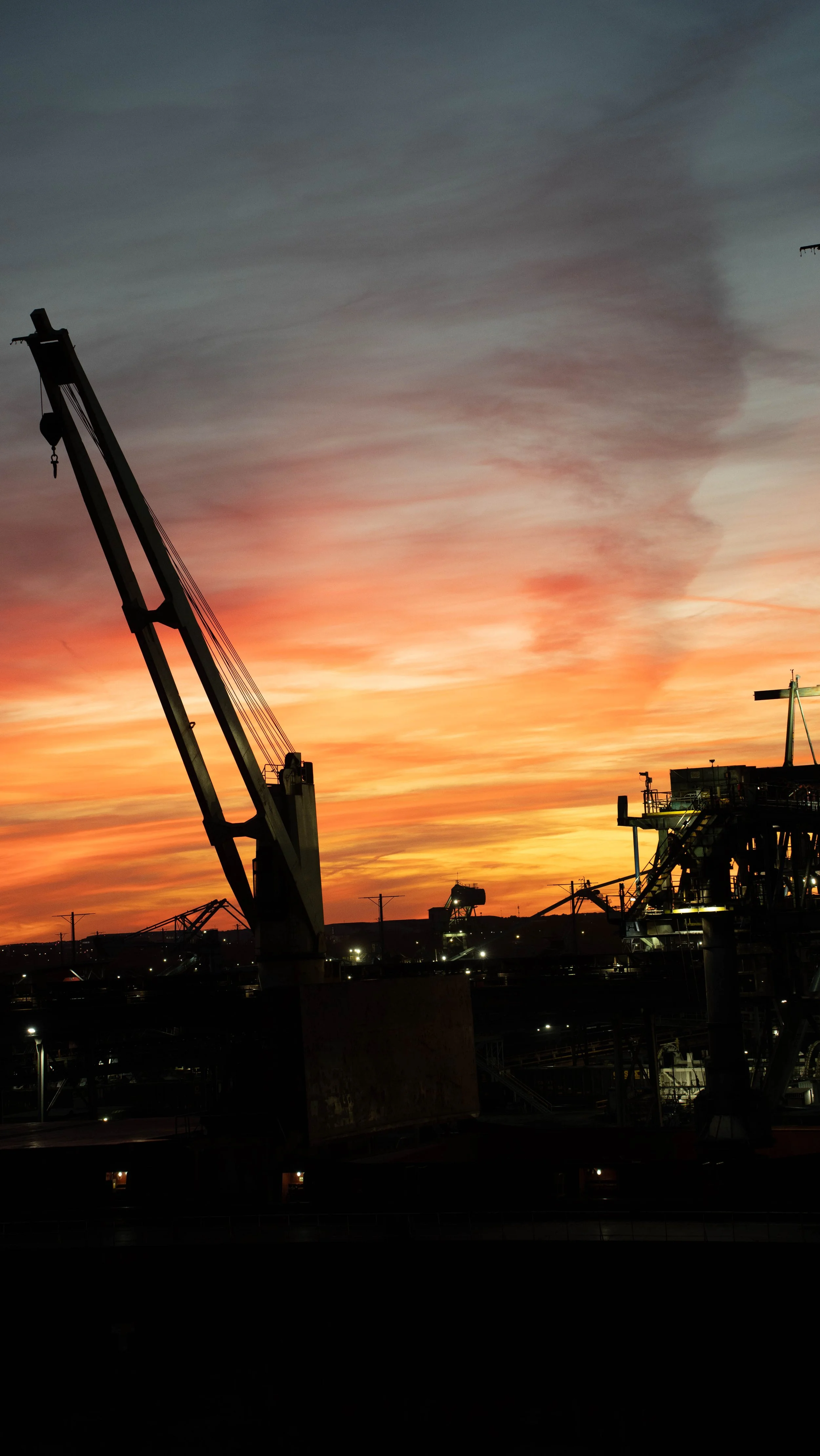 Silhouetted cranes and industrial structures against a colorful sunset in a port or construction site.