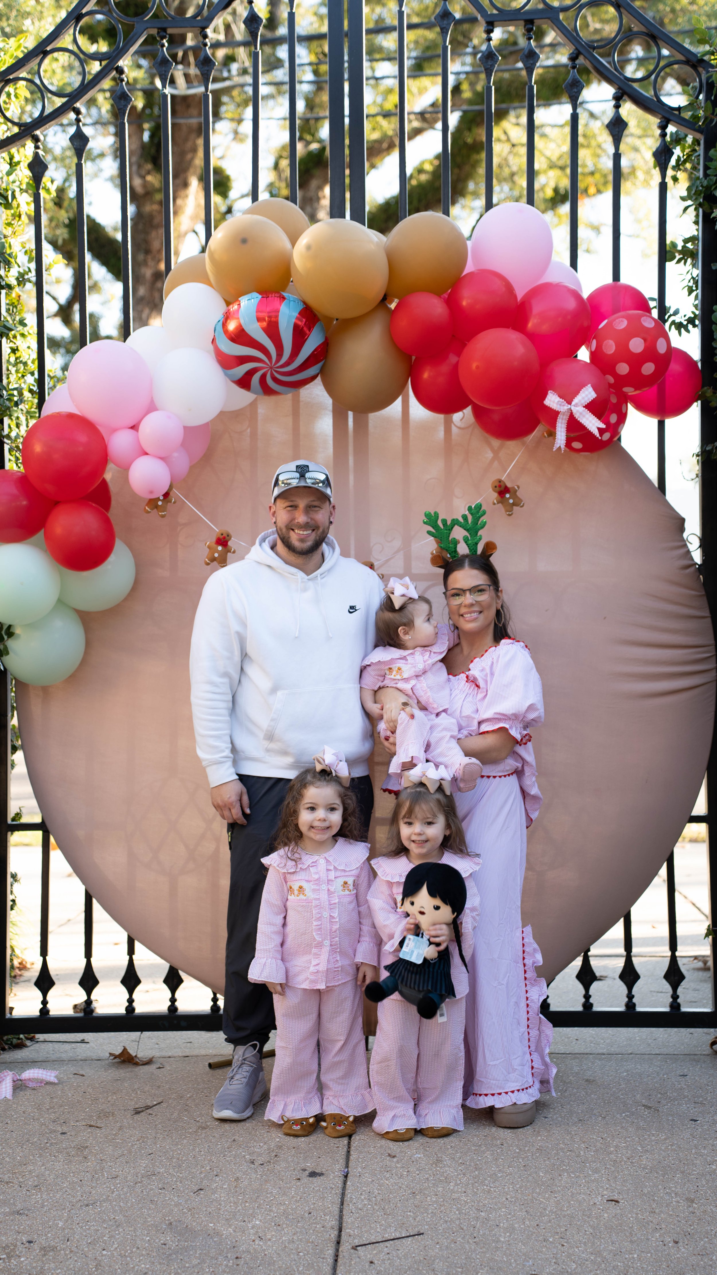 A family of five, including two adults and three young girls, poses in front of a large peach-shaped backdrop decorated with colorful balloons and holiday-themed ornaments. The children are dressed in pink pajamas, and two of the girls wear bunny ear headbands. The woman has reindeer antler headbands, and one of the girls holds a doll. They are smiling outdoors on a sunny day.
