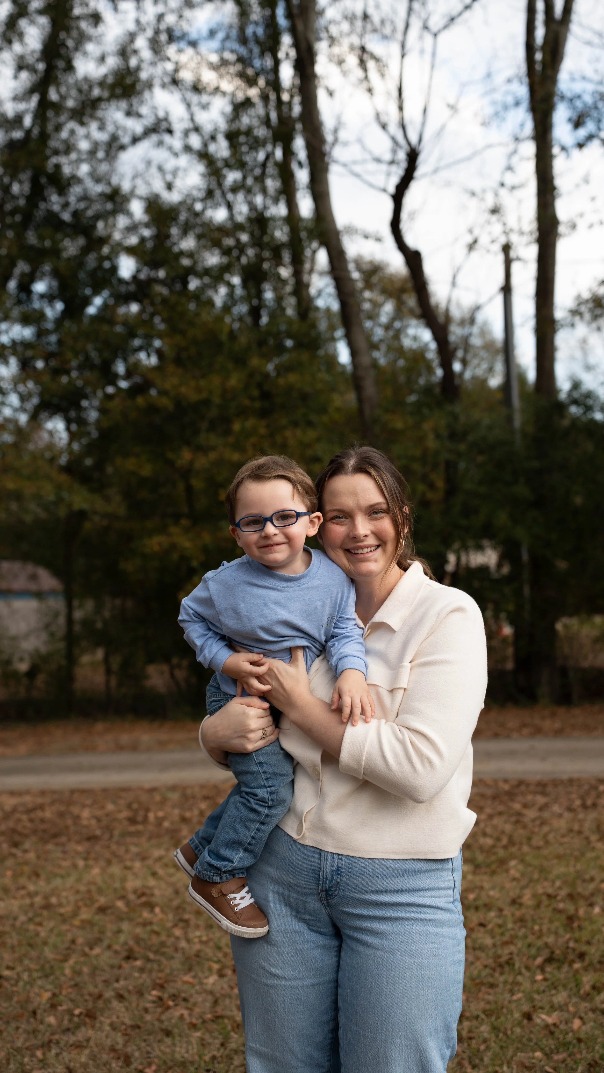 A woman holding a young boy with glasses outdoors, trees and cloudy sky in the background.