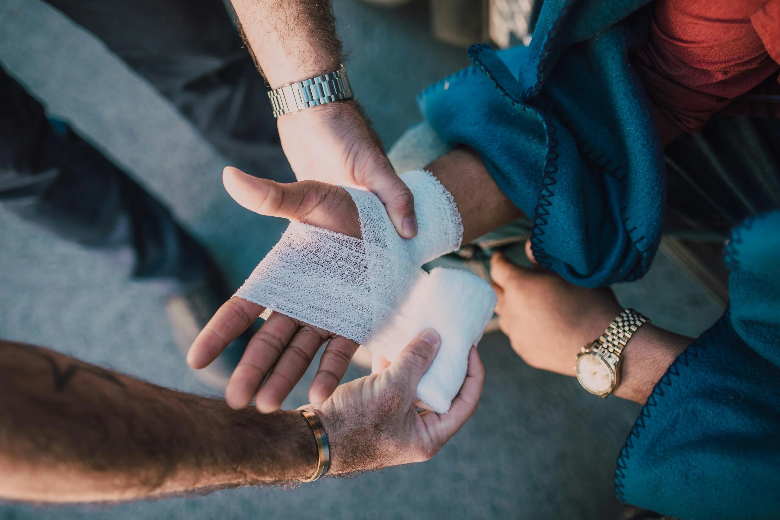Two people helping an injured person with a hand wrapped in bandages, and one of the helpers is holding the injured person's hand, in an outdoor setting.