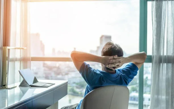 A man sitting on a chair facing a large window, looking out at a cityscape with his hands behind his head. There is a laptop on the desk beside him.