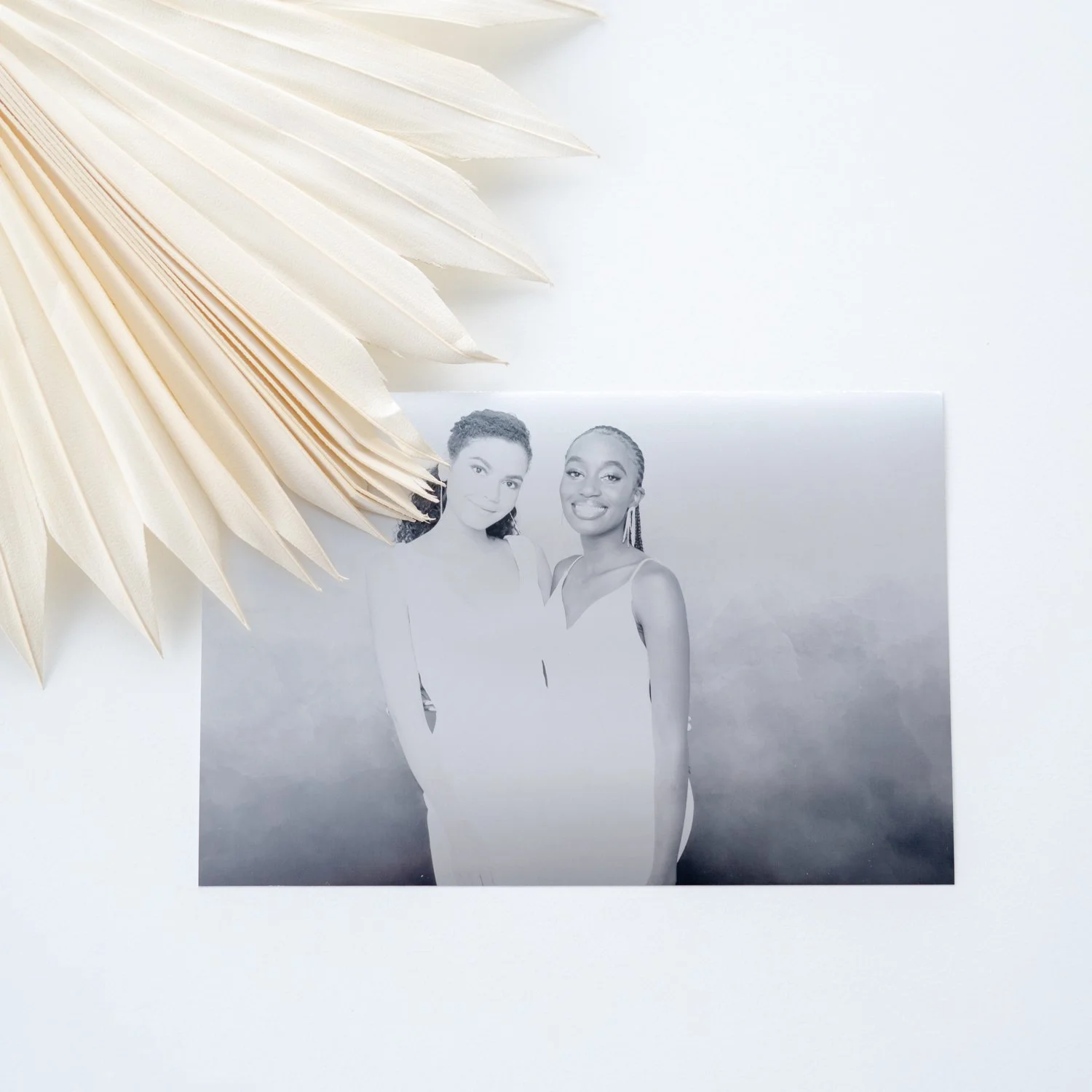 Black and white photo of two women standing outdoors, both smiling, with one wearing a sleeveless dress and the other a deconstructed sleeveless top, with a cloudy sky background.