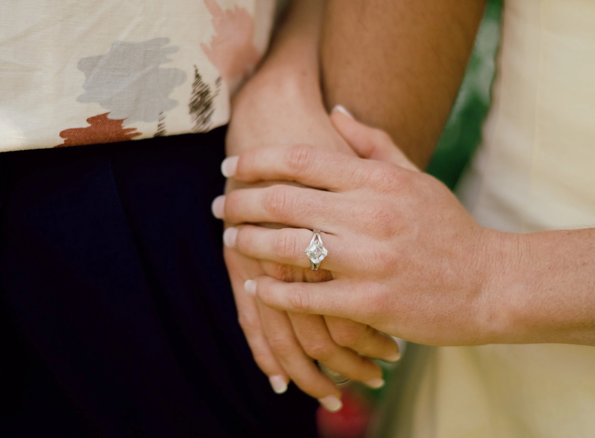 Close-up of a woman's hand with a diamond engagement ring, gently resting on a man's torso, with their hands linked.