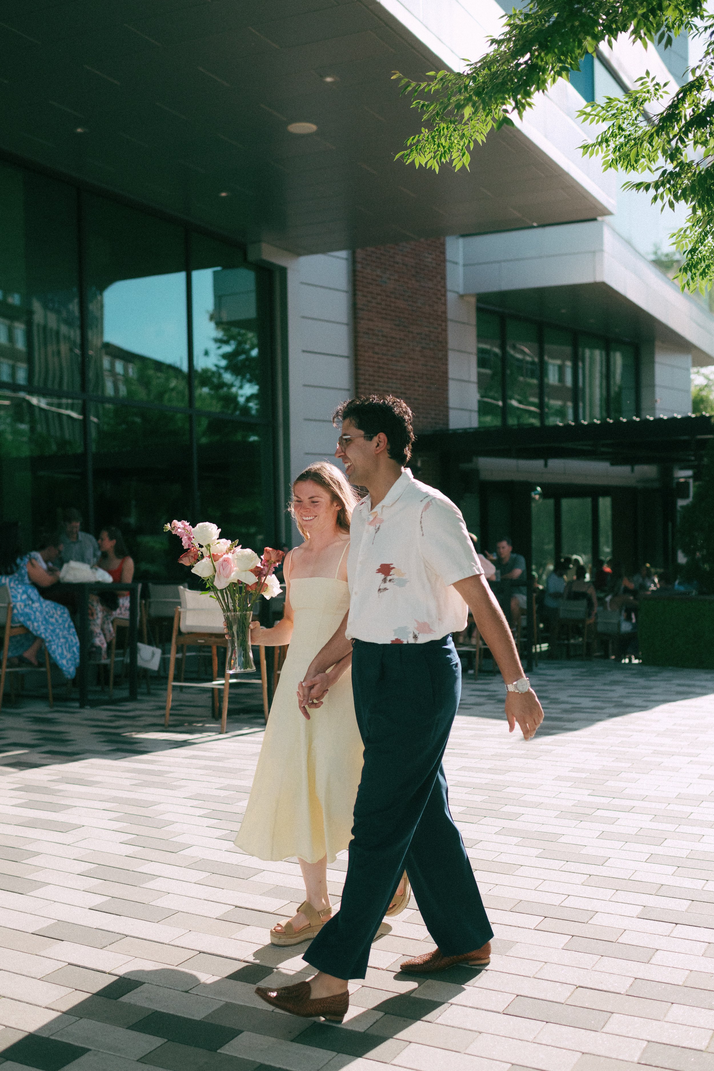 A young couple walks hand in hand outside a modern building on a sunny day. The woman is in a yellow dress holding a bouquet of flowers, and the man is in a white shirt with a pattern and dark pants. They are smiling and seem happy.
