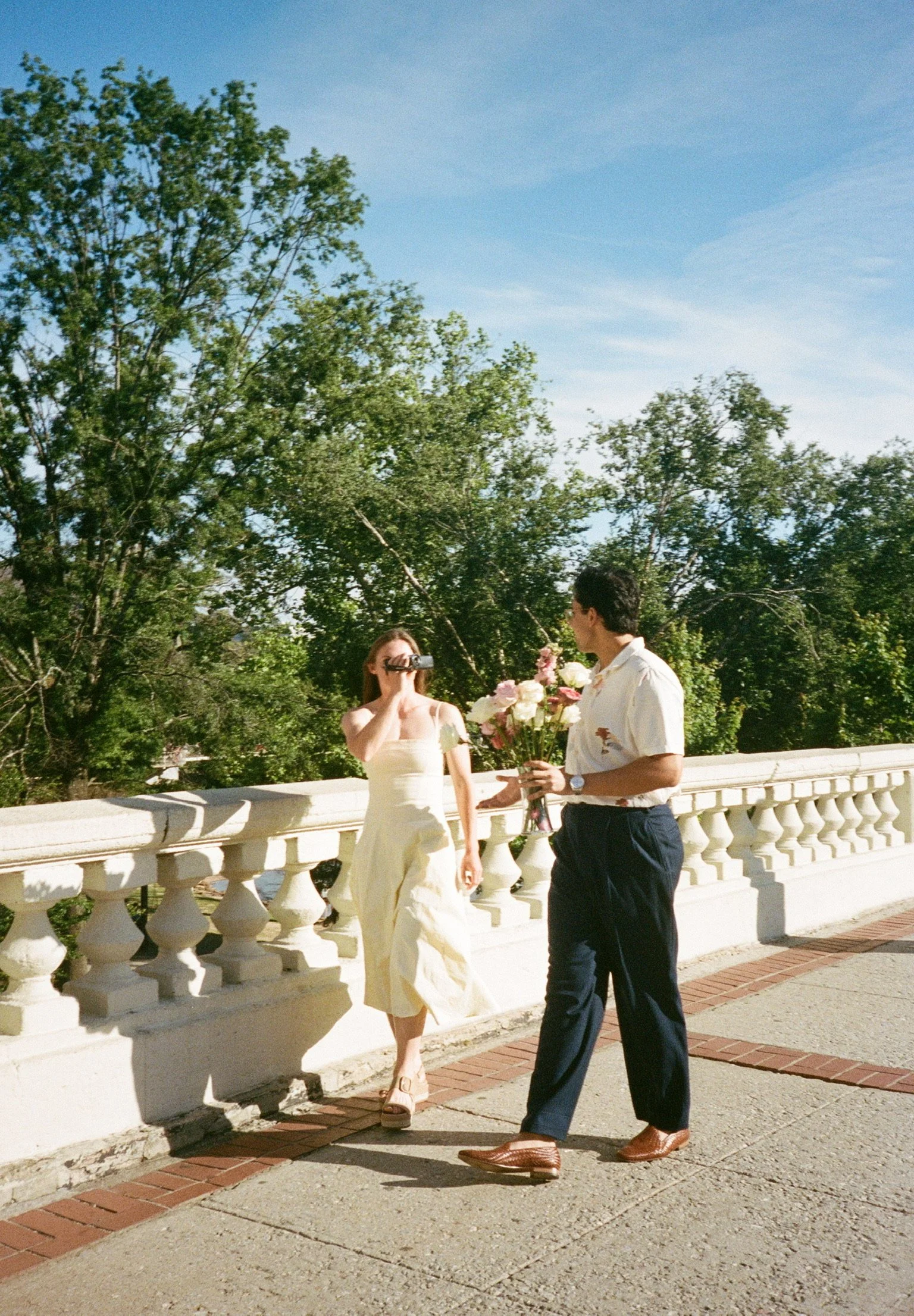 A woman in a white dress taking a photo with a camera while a man in dark pants and a white shirt offers her a bouquet of pink and white roses on a bridge with trees in the background.
