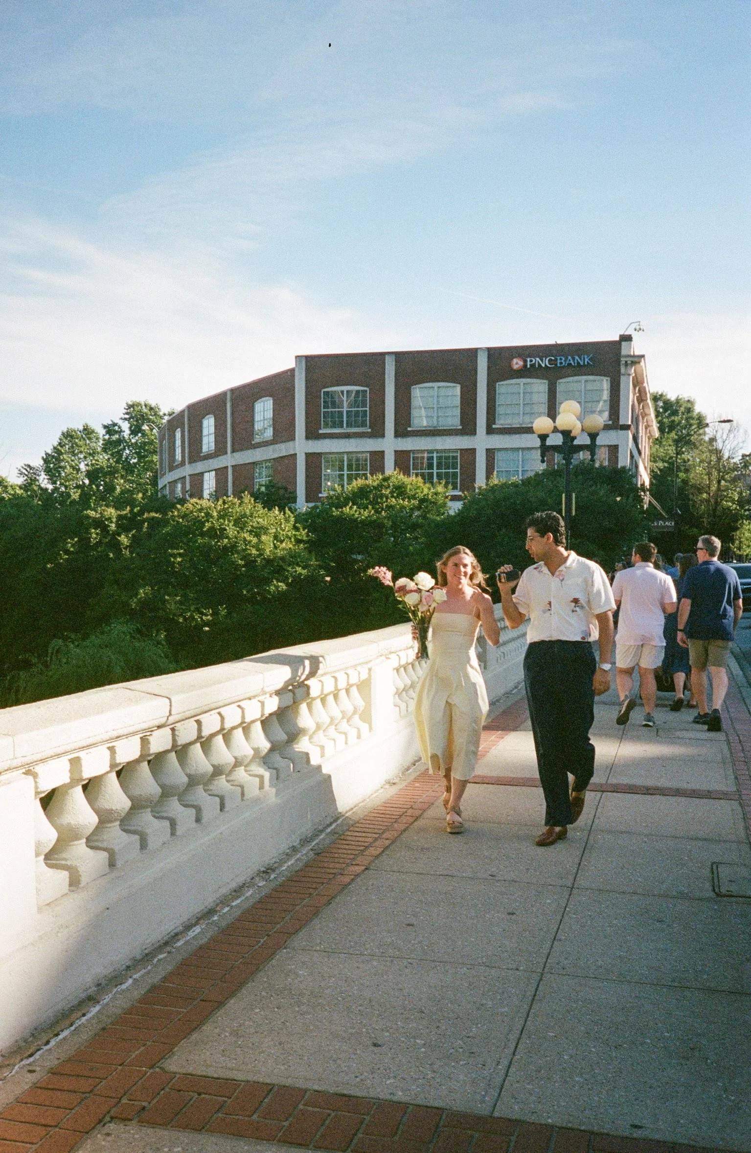A couple in wedding attire walking on a bridge, with the bride holding a bouquet of pink and white flowers and the groom holding a microphone, as other people walk behind them