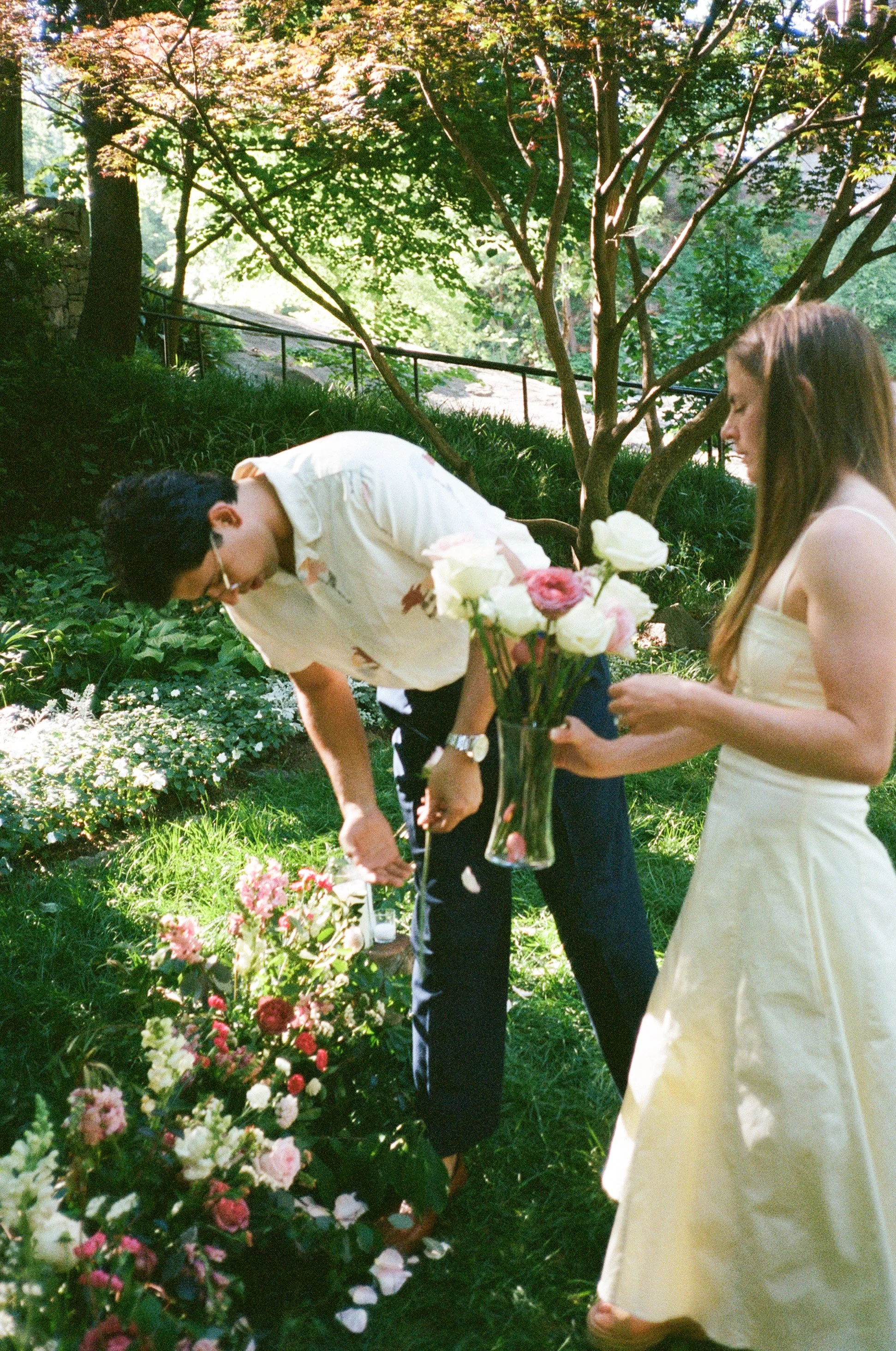 A man and a woman planting flowers in a garden, with the woman holding a vase of flowers and the man using a gardening tool, surrounded by lush greenery and trees on a sunny day.