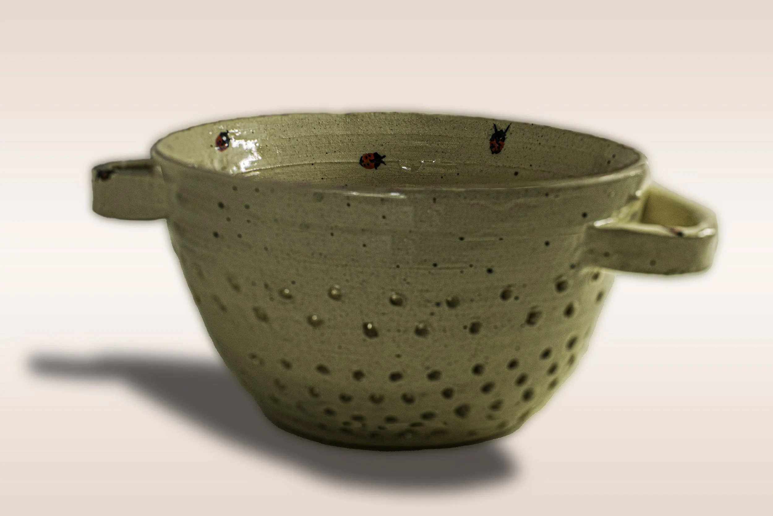 Cream-colored ceramic colander with small holes and two handles, decorated with red and black ladybugs, placed against a plain background.