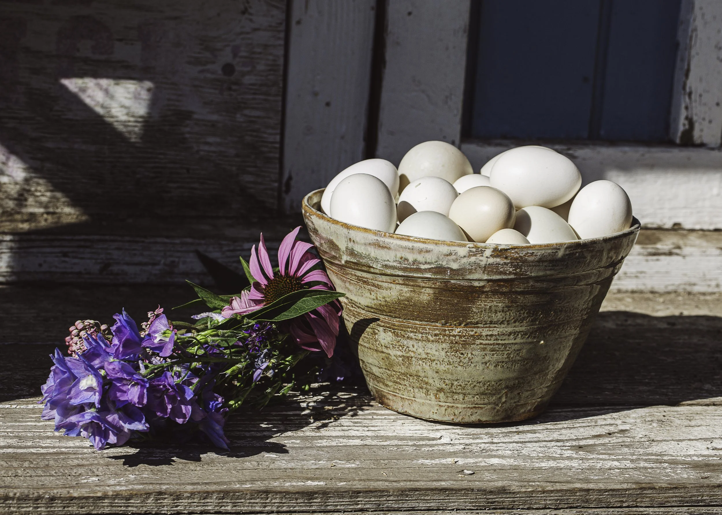 A rustic ceramic bowl filled with white eggs sitting on a weathered wooden surface, next to a small bouquet of colorful flowers including purple and pink blooms.