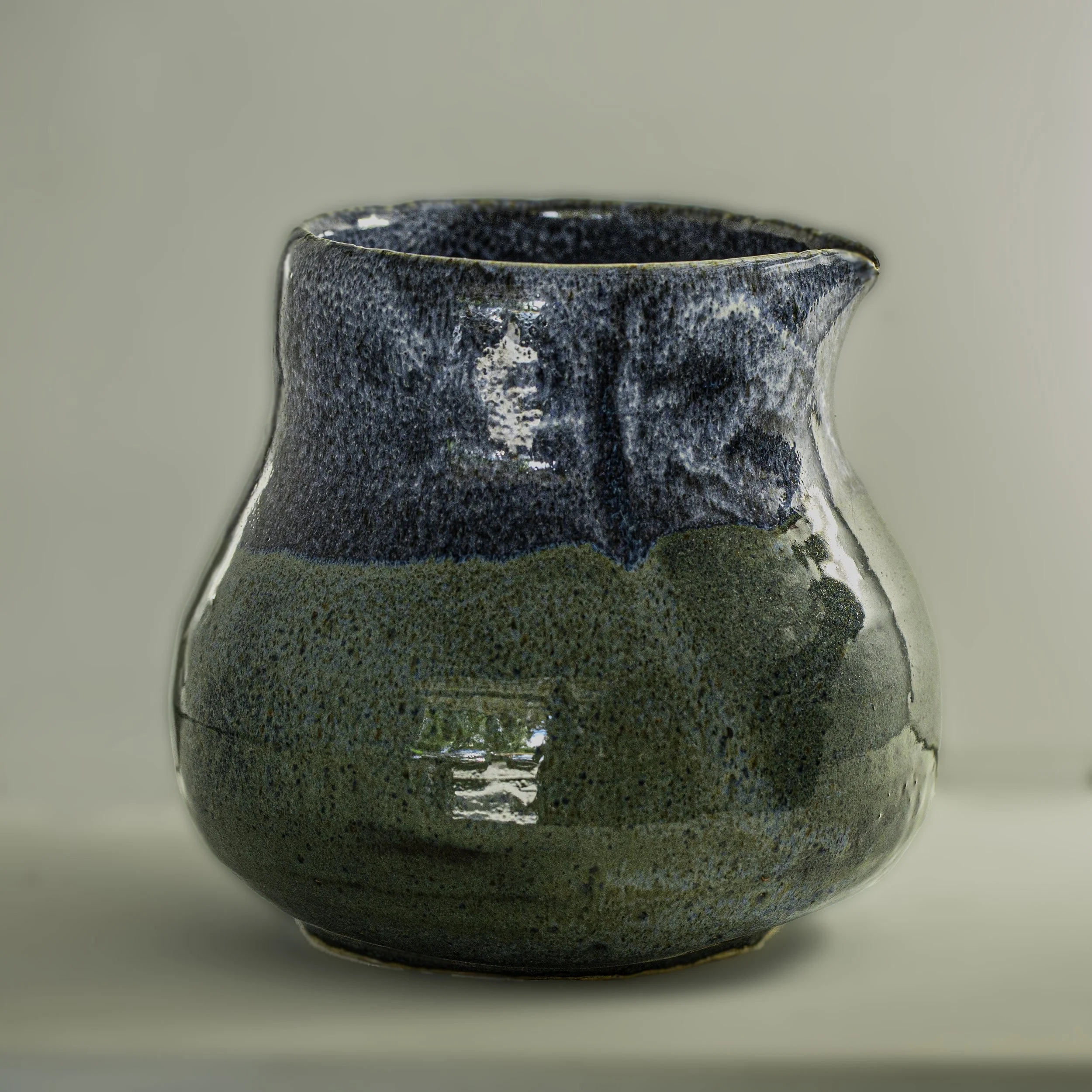 A ceramic teapot with a textured, glazed surface in shades of blue and green, sitting on a light-colored surface against a plain background.