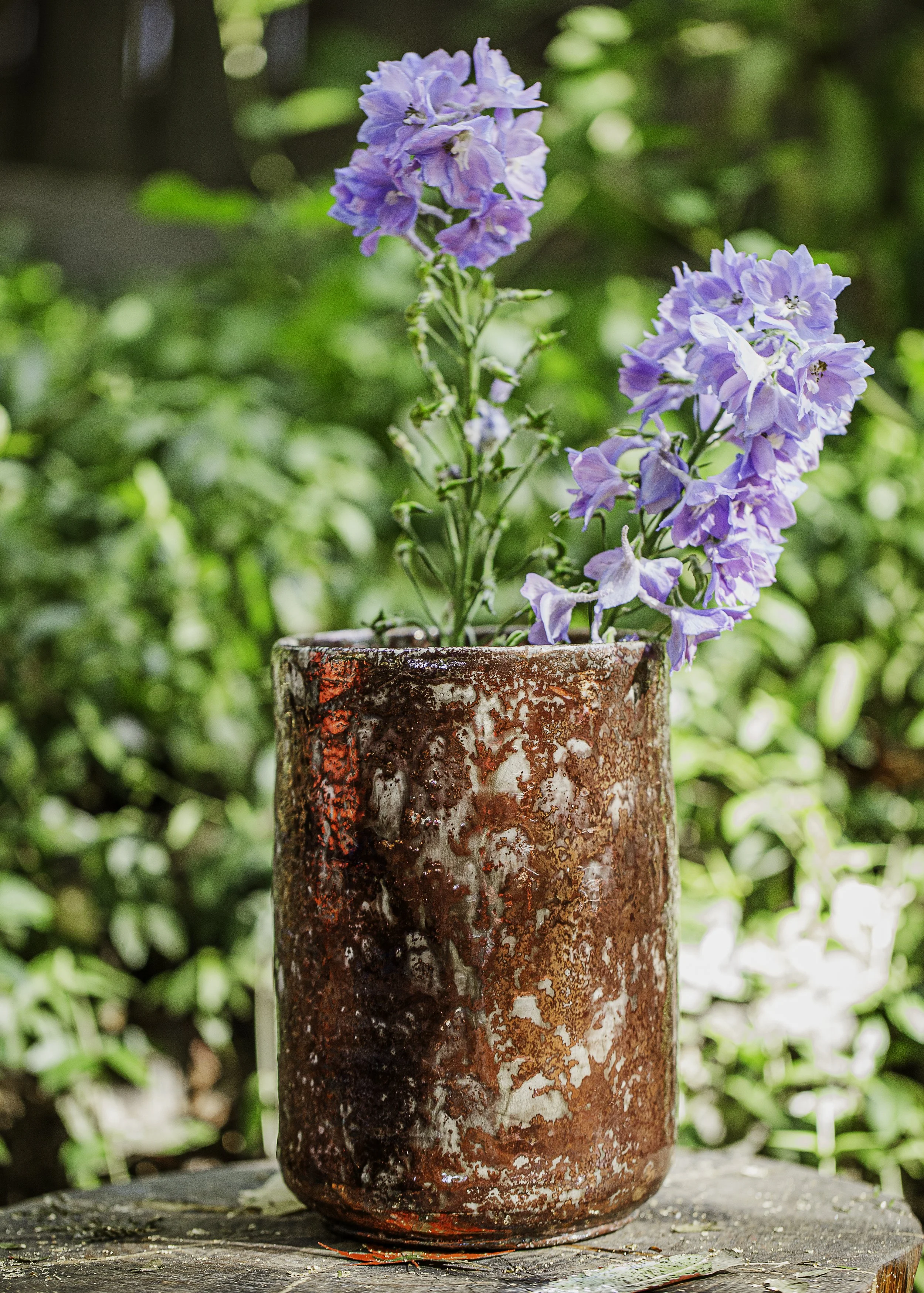 A tall, cylindrical, rusty red ceramic vase with a textured surface, holding a purple flower with multiple blossoms, placed outdoors on a wooden surface with a green leafy background.