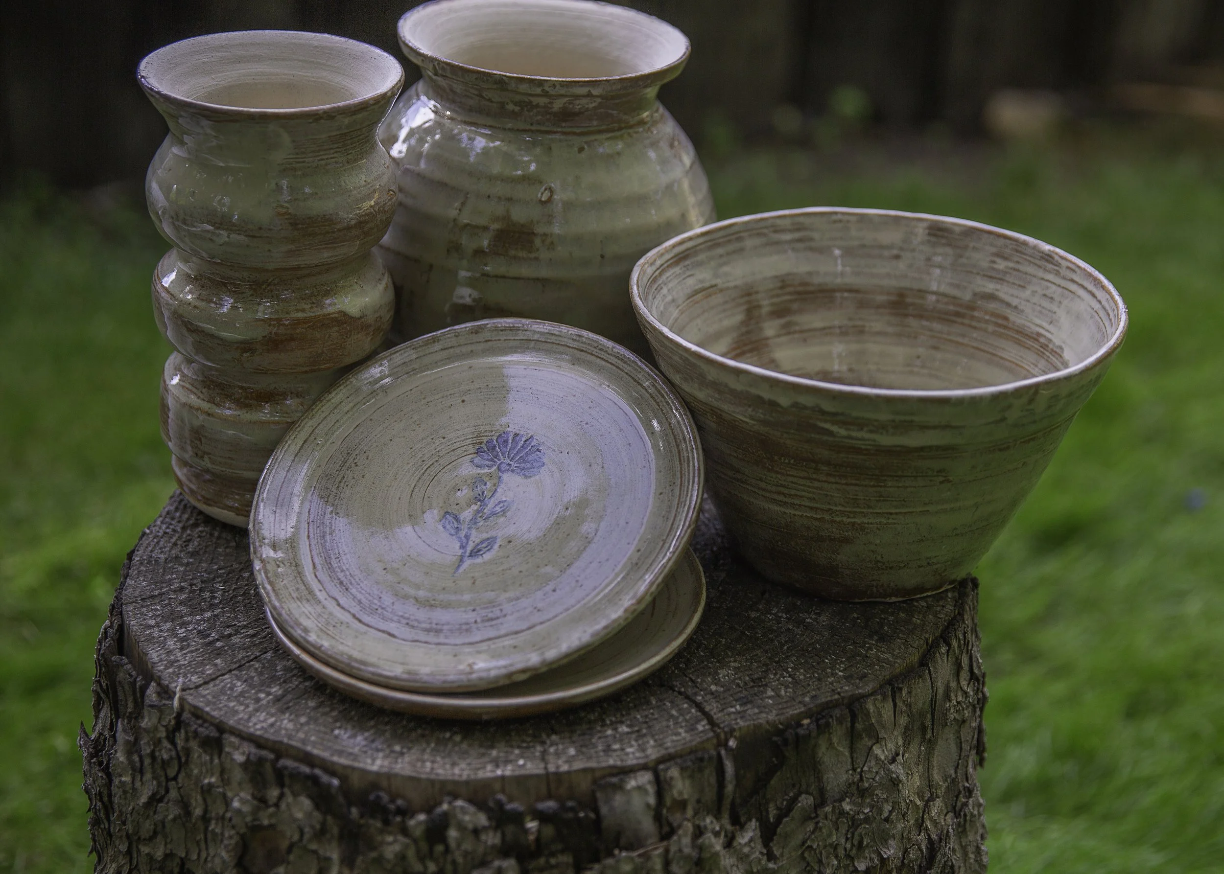 Set of handmade ceramic bowls and plates with earthy glaze, placed on a tree stump outdoors, with a grassy background.
