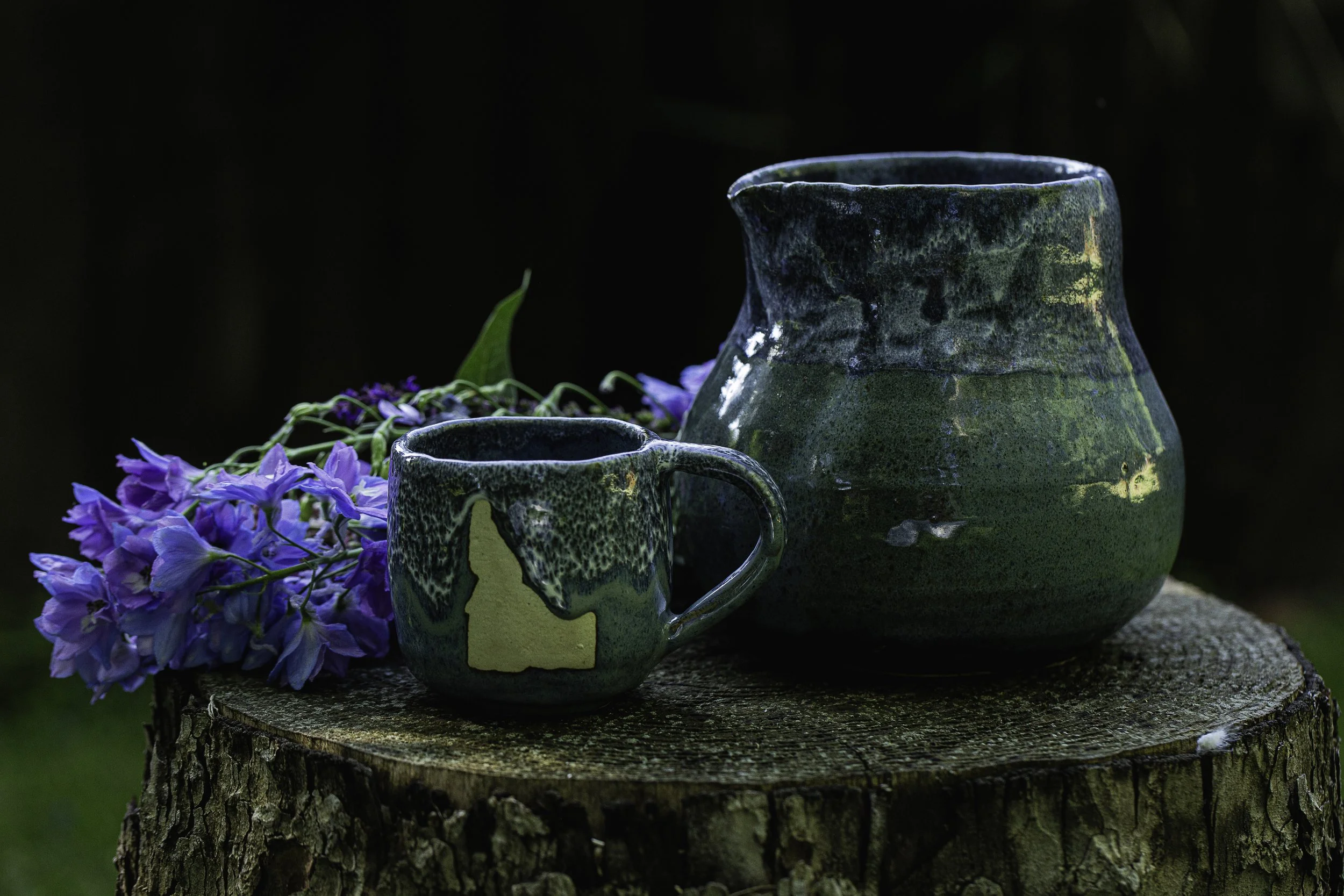 A green ceramic mug with a large handle and a larger matching pitcher sit on a tree stump, with purple flowers lying beside them against a dark background.