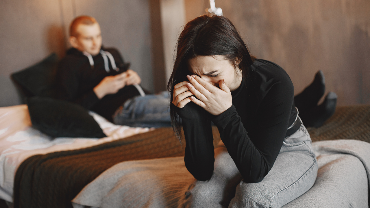 A woman sitting on a bed with her head in her hands, visibly upset, while a young man in the background is lying on the bed, looking at his phone.