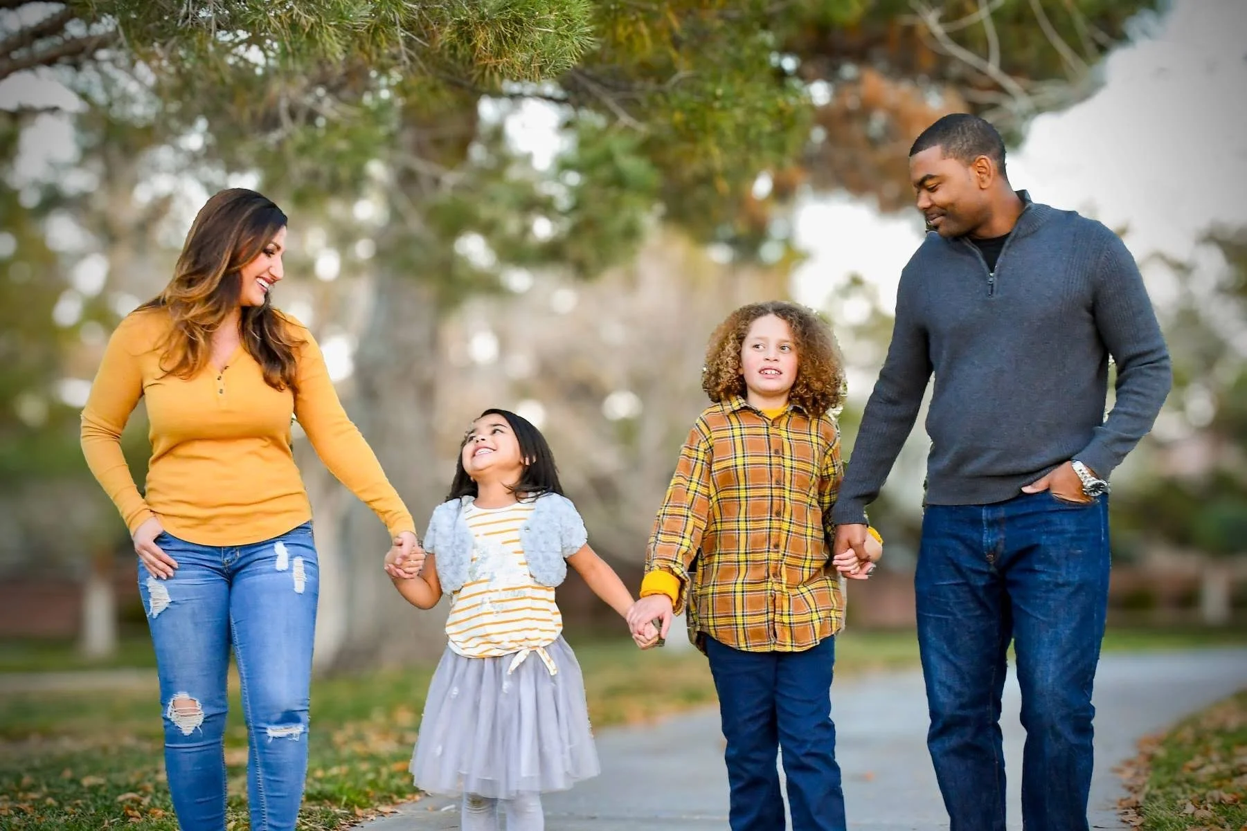 A multiracial family of four walking outdoors in a park, holding hands, and smiling.