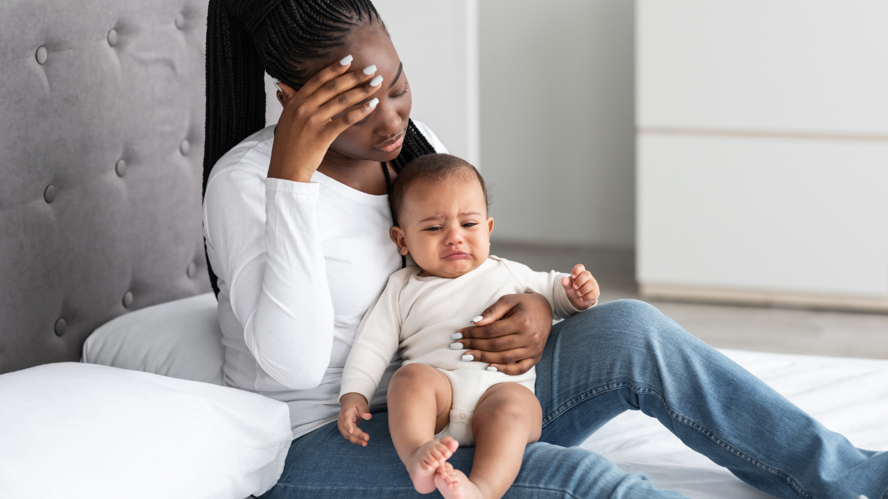 An African American woman sitting on a bed, holding a crying toddler on her lap, with one hand on her forehead, appearing distressed.
