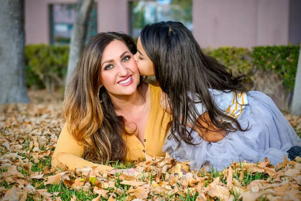 A woman and a young girl lying on the ground covered in fallen leaves, with the girl giving the woman a kiss on the cheek. The woman has long, wavy brown hair and is smiling, wearing a yellow top. The girl has long dark hair, wearing a gray and white dress with yellow stripes.