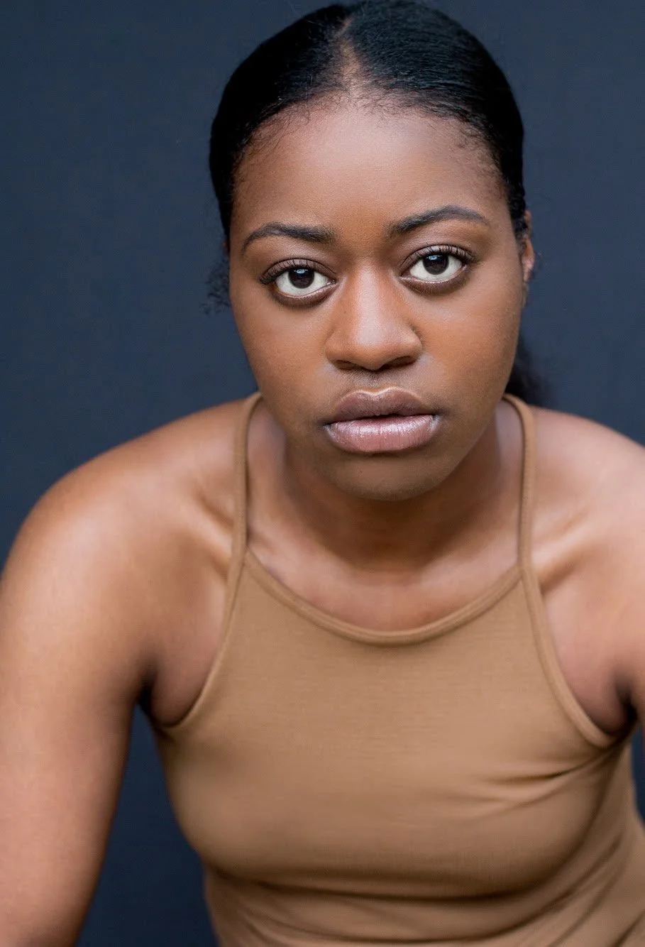 A young woman with dark hair pulled back, wearing a tan tank top, looking directly at the camera with a serious expression against a dark background.