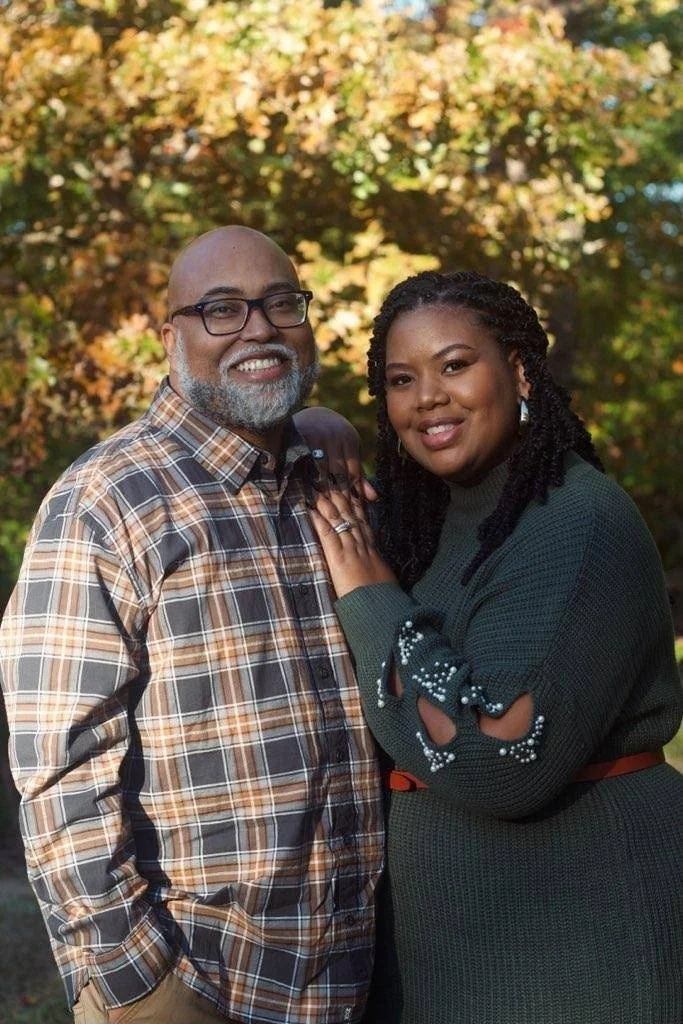 A smiling man with glasses and a beard wearing a plaid shirt, and a woman with braided hair wearing a dark green sweater, standing outdoors in front of trees with fall foliage.