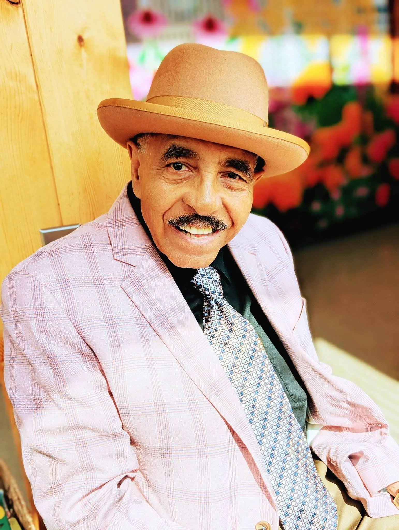 A smiling man wearing a pink plaid blazer, a patterned tie, a black shirt, and a tan cowboy hat.