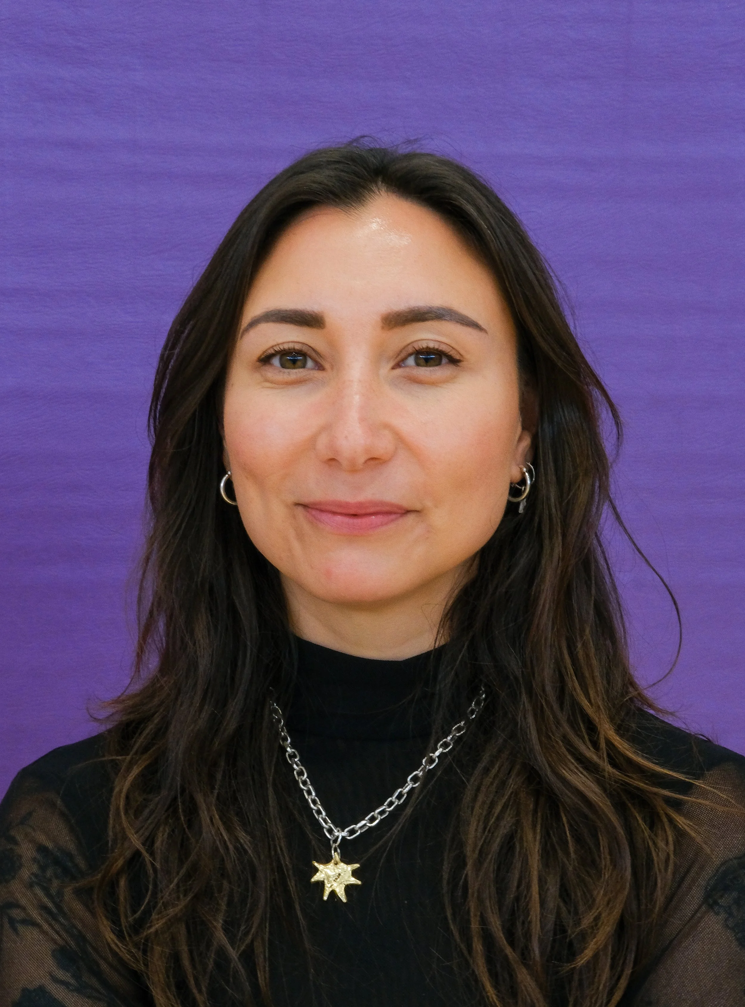 Close-up of a woman with long brown curly hair, wearing a leopard print top, a necklace with a cross pendant, and a chain with a pendant, standing against a plain off-white wall.