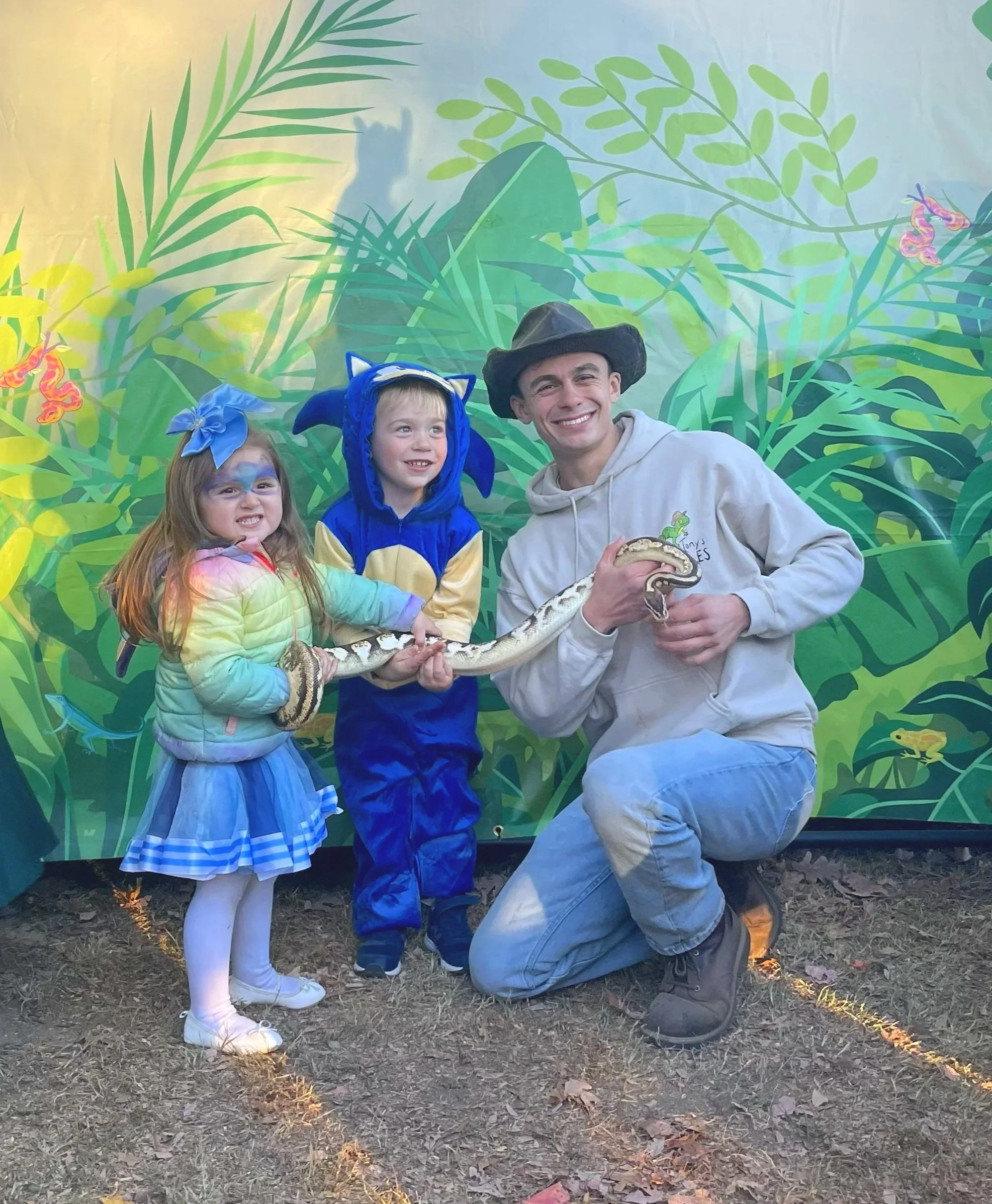 A man and two young girls posing with a snake in front of a jungle-themed backdrop, outdoors on a dirt ground. The best reptile show in NY