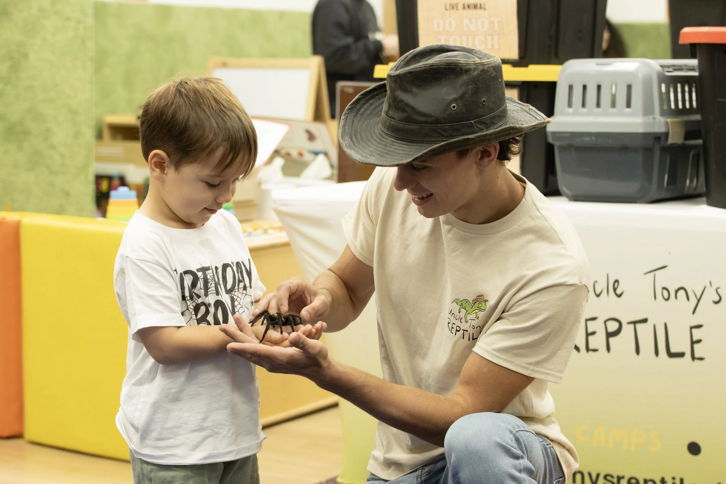 A young boy and an adult man are examining a large spider together at an indoor event. The man is wearing a beige t-shirt with a logo and a wide-brimmed hat, while the boy is wearing a white t-shirt with black text. They are smiling and looking at the spider.