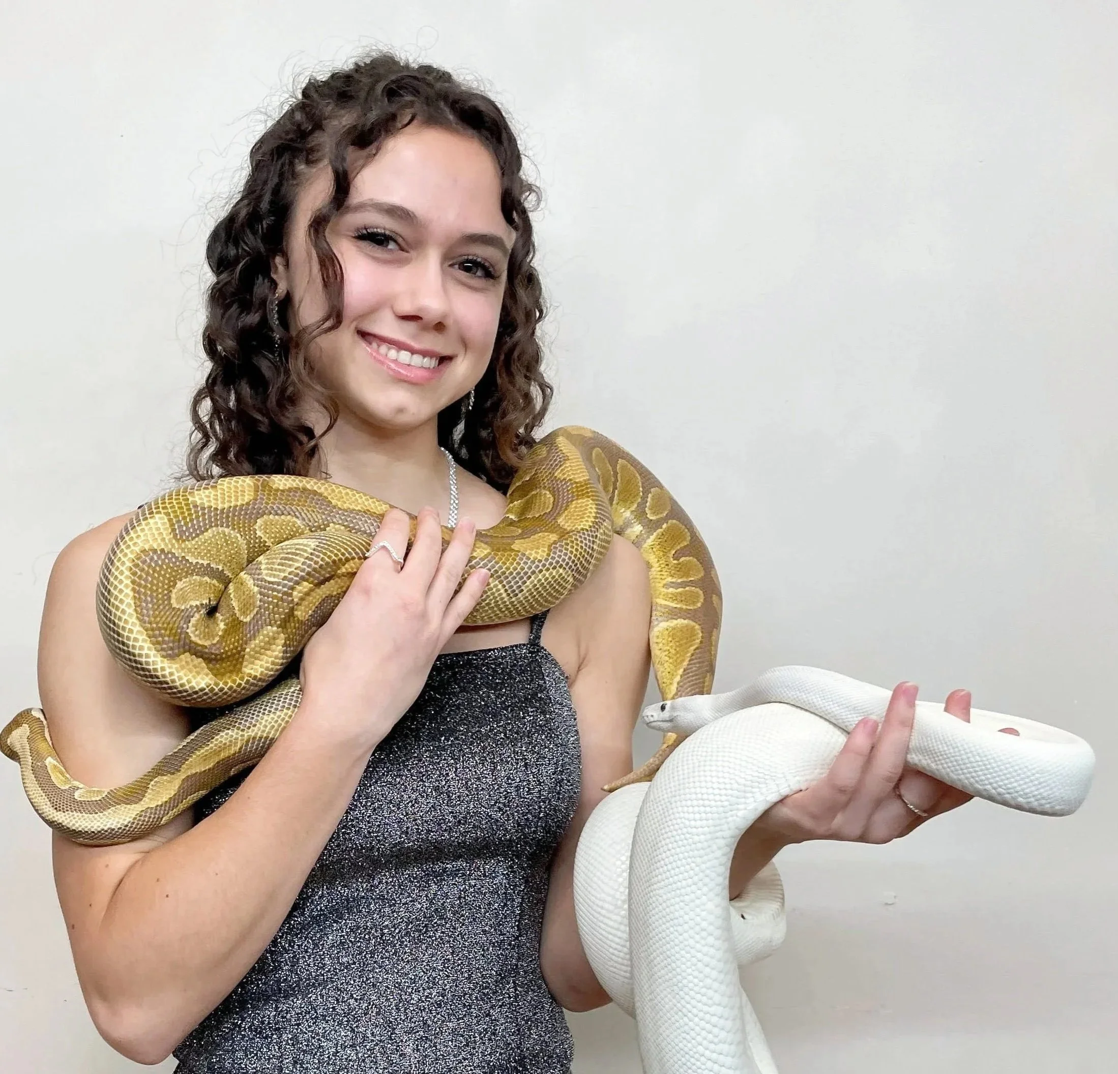 Young woman with curly brown hair smiling while holding a white snake around her shoulders, standing against a plain white wall.