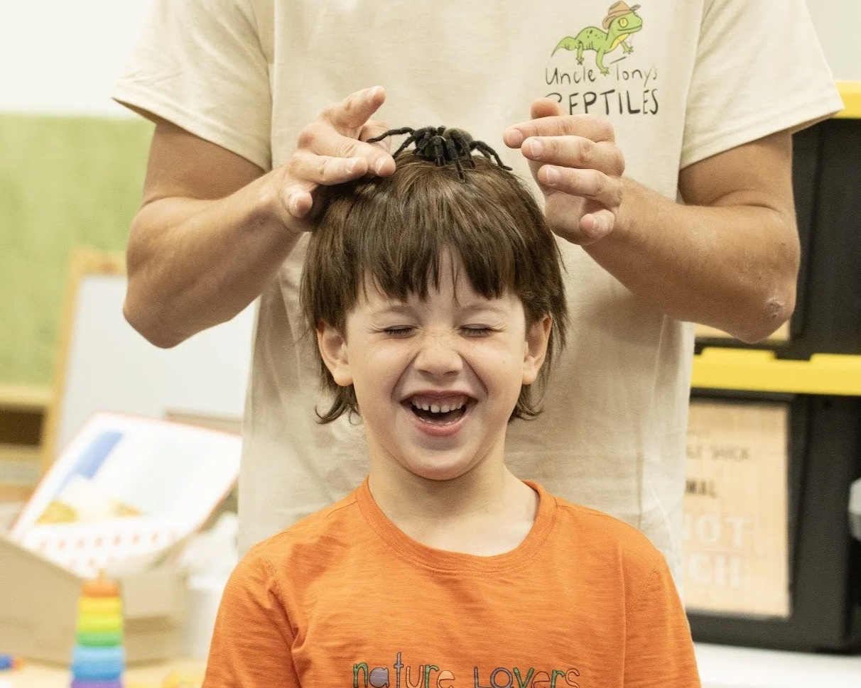 A boy laughing while someone is placing a tarantula on his head in a classroom or educational setting.