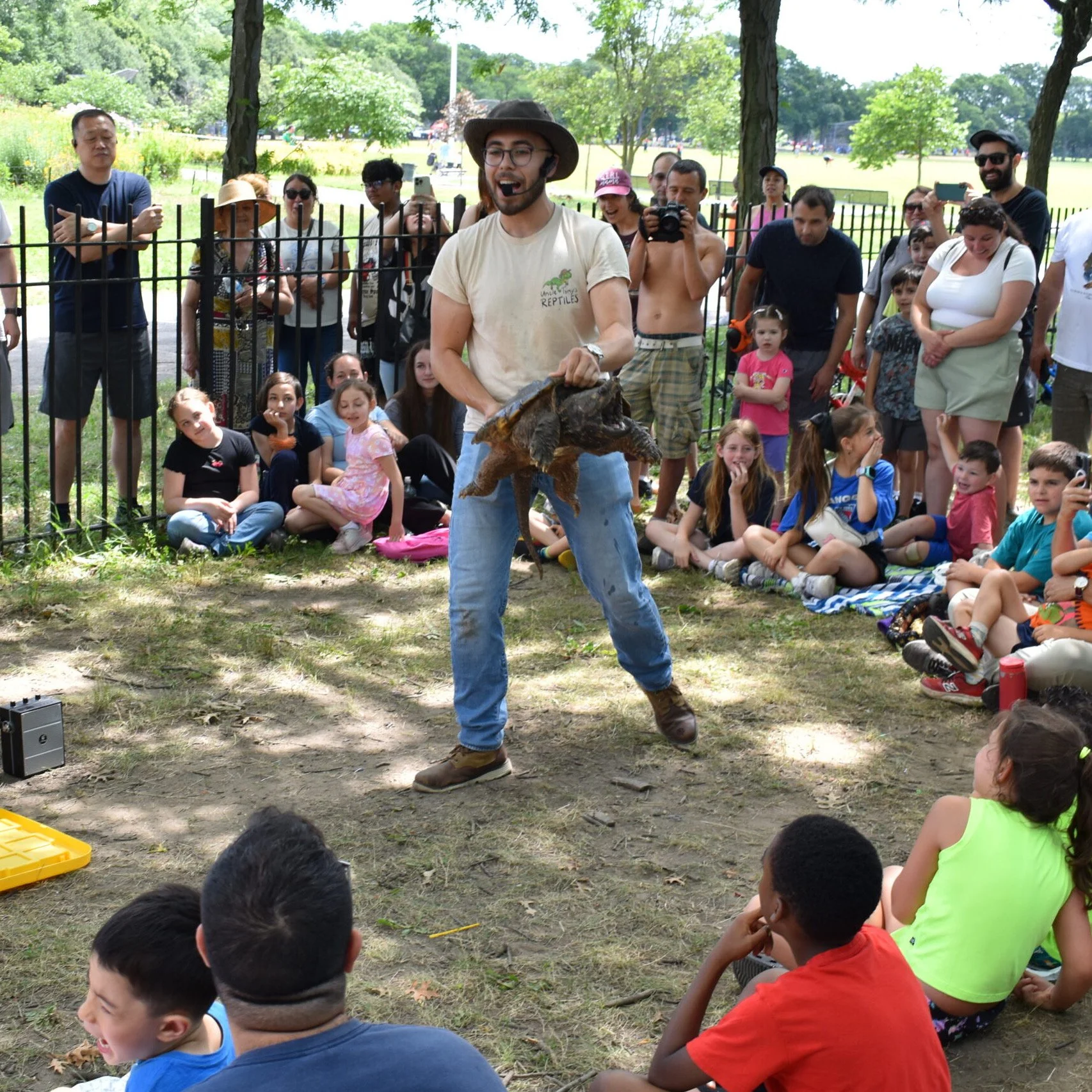 A man in a beige T-shirt, jeans, and a wide-brimmed hat holding a large turtle in front of an audience of children and adults at a park, with trees and open grassy areas in the background.