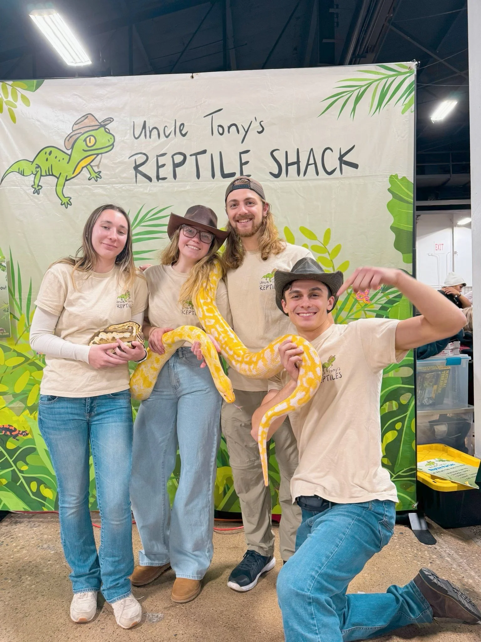 Four people posing with a large yellow and white snake in front of a backdrop that reads 'Uncle Tony's Reptile Shack' decorated with cartoon reptiles and jungle leaves.
