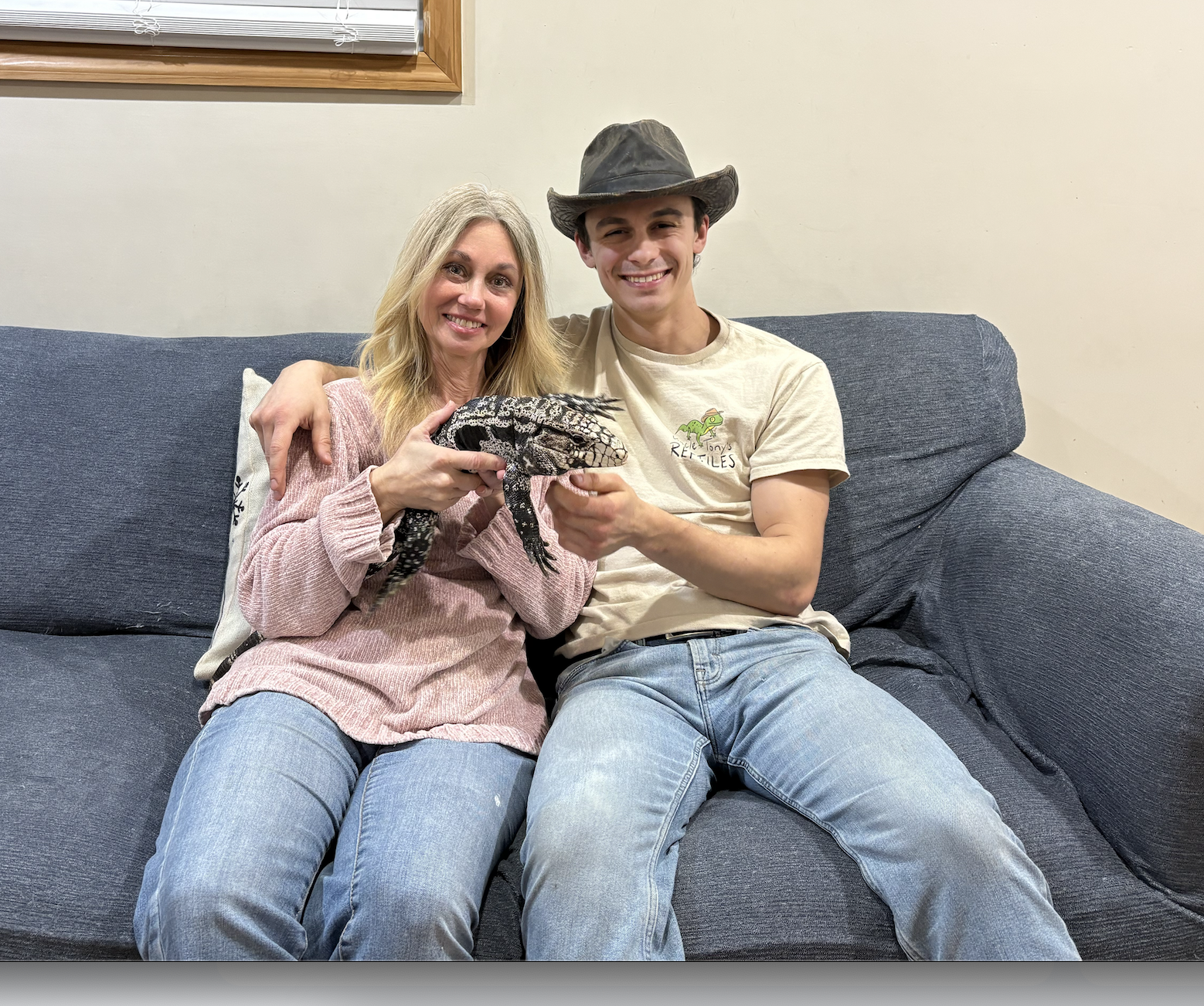 A woman and a young man sit on a navy blue couch, smiling at the camera. The woman is holding an Argentine black and white tegu lizard, and the young man is wearing a wide-brimmed hat and a beige T-shirt with a chameleon logo. They are indoors with a window and beige wall in the background.