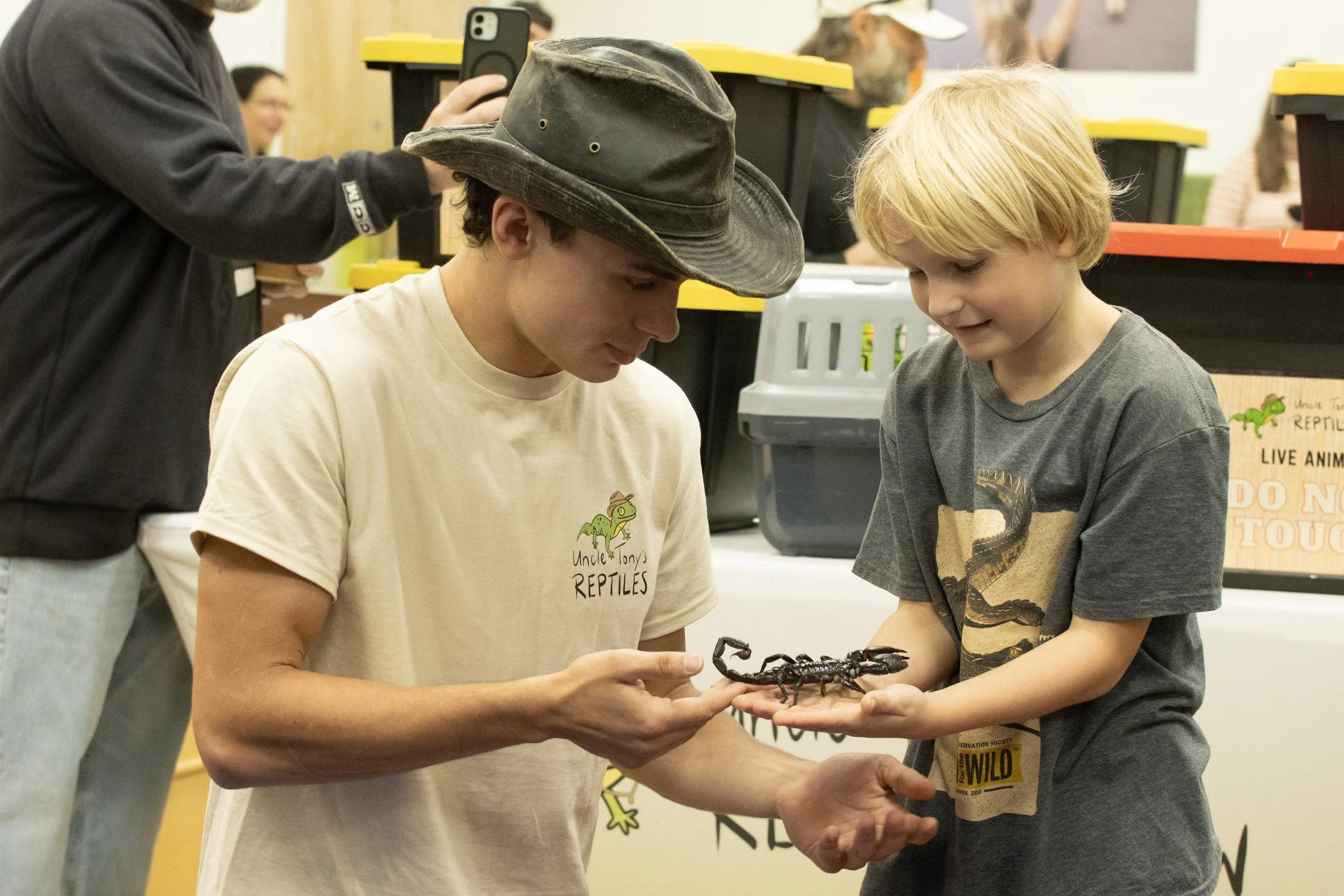 A young man and boy examining a scorpion at Uncle Tony's Reptiles booth during an indoor summer camp event.